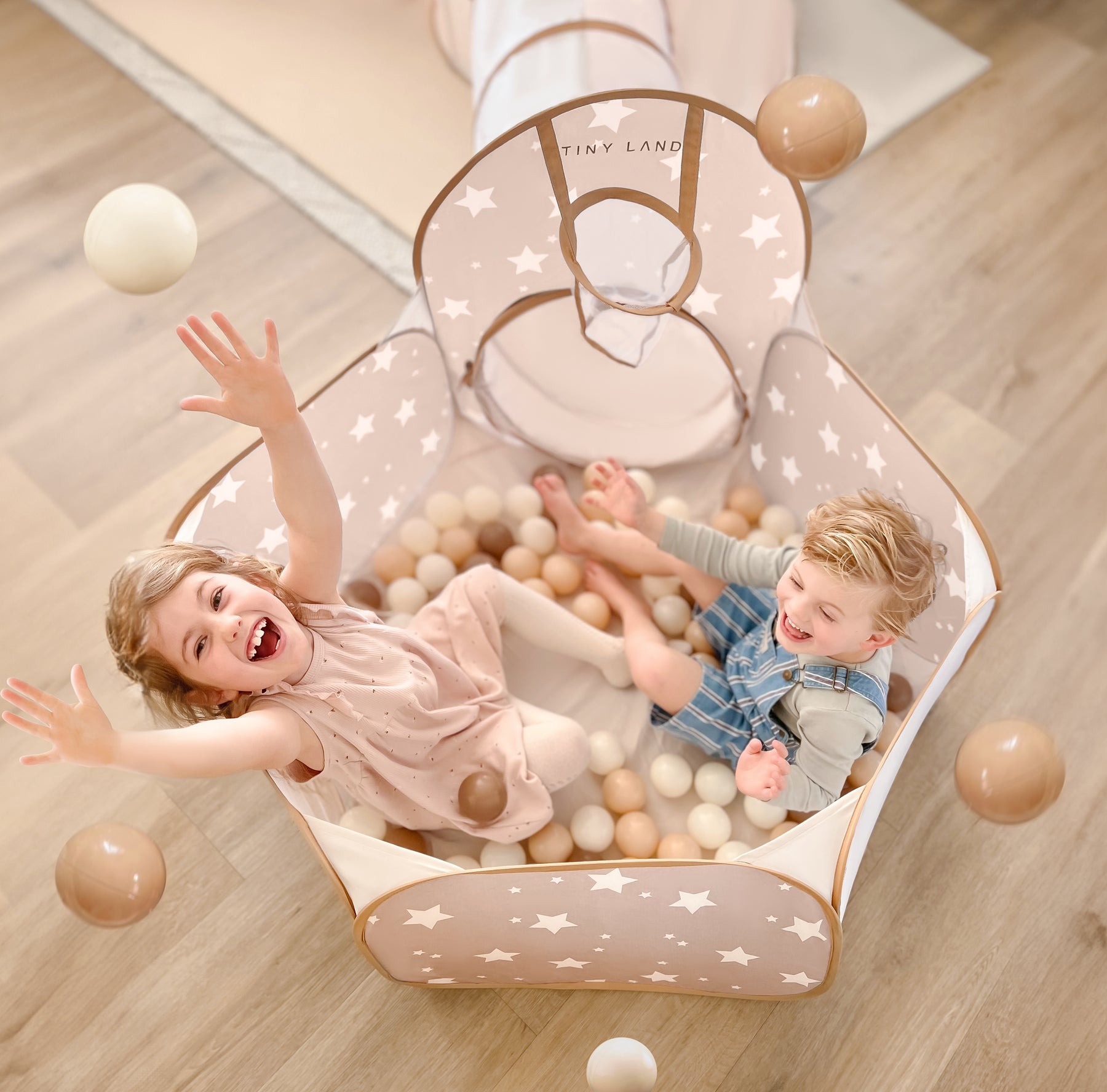 Two young children laugh and play in a beige playpen with white stars, tossing Tiny Land® Ocean Ball - 200 pcs with Varied Colors by Tiny Land. The playpen is on a light wood floor, featuring a soft mat and an attached tunnel.