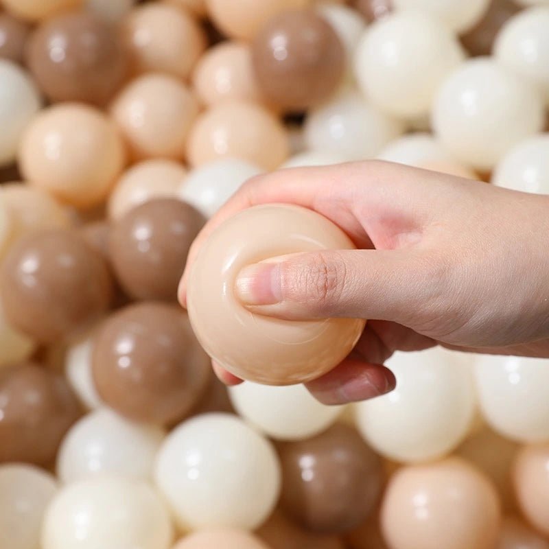 A hand squeezes a soft beige ball from the Tiny Land® Discovery 5-in-1 Play Tunnel & Ocean Ball Set, surrounded by smooth ocean balls in shades of beige, brown, and white.