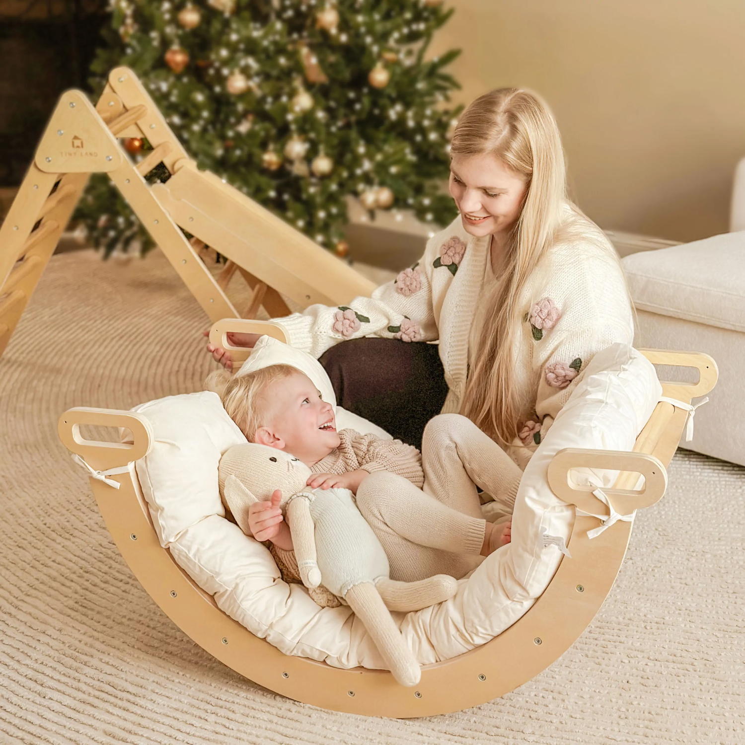 A woman and child sit in a wooden rocker with the Tiny Land® Thick Padded Play Cushion. The child holds a stuffed toy, and a decorated Christmas tree glows in the background as they smile at each other.