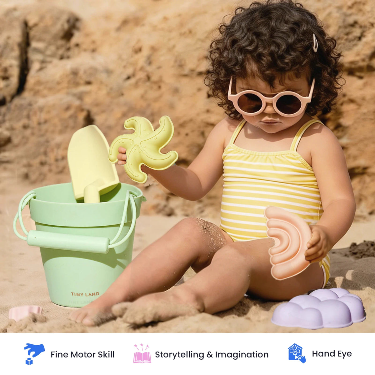 A young child in a yellow striped swimsuit and round sunglasses engages in sensory play with the Tiny Land® Sand-Buddies Sand Toy Set by Tiny Land—including a bucket, shovel, and pastel sand molds—while sitting on sandy ground near rocks.