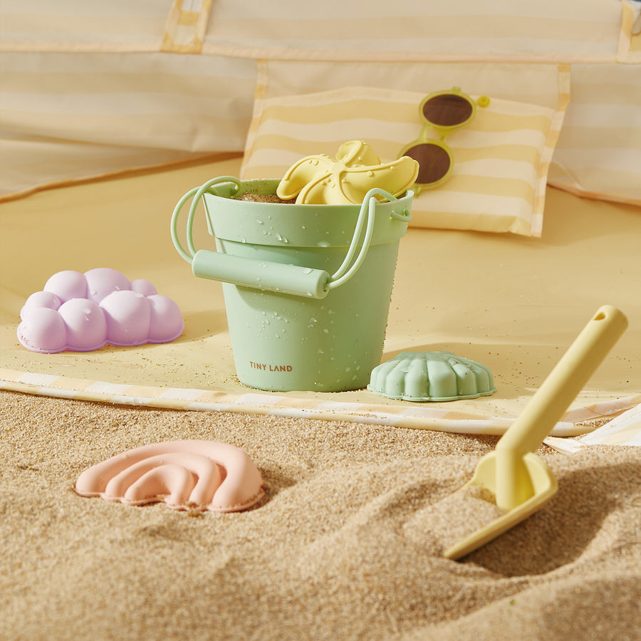 A pastel green bucket from the Tiny Land® Sand-Buddies Sand Toy Set sits on the beach with pastel rainbow and cloud molds, plus a yellow spade. A striped pillow and sunglasses are visible in the background.