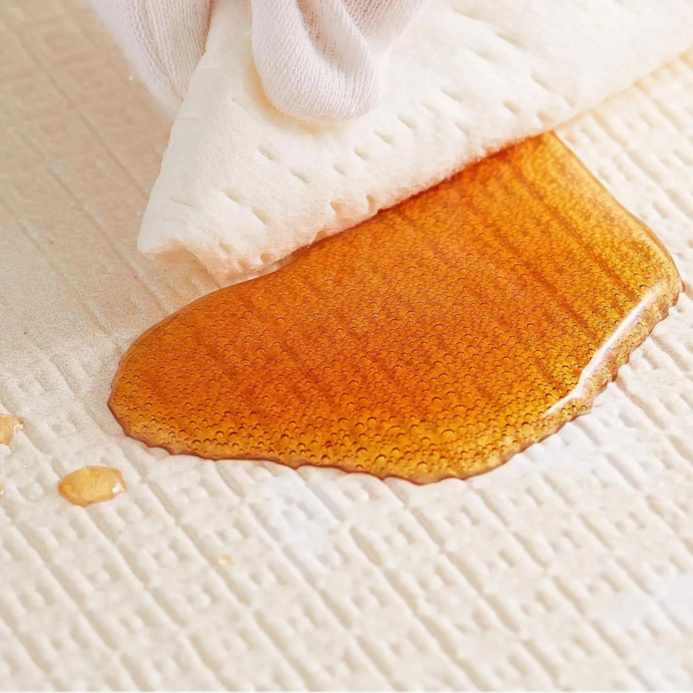 A white-gloved hand uses a cloth to blot up a large amber liquid spill on the textured, waterproof surface of the Tiny Land® Double-Sided Baby Playmat Forest Track Wonder by Tiny Land.