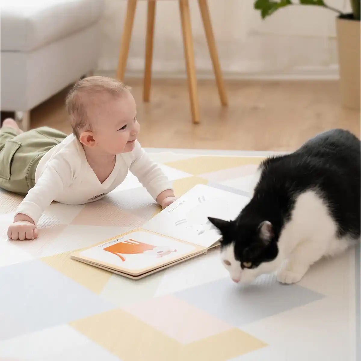 A smiling baby relaxes on the Tiny Land® Double-Sided Baby Playmat Urban Zoo Adventure by Tiny Land, next to an open book, watching a black and white cat explore the playmat in a bright, cozy room.