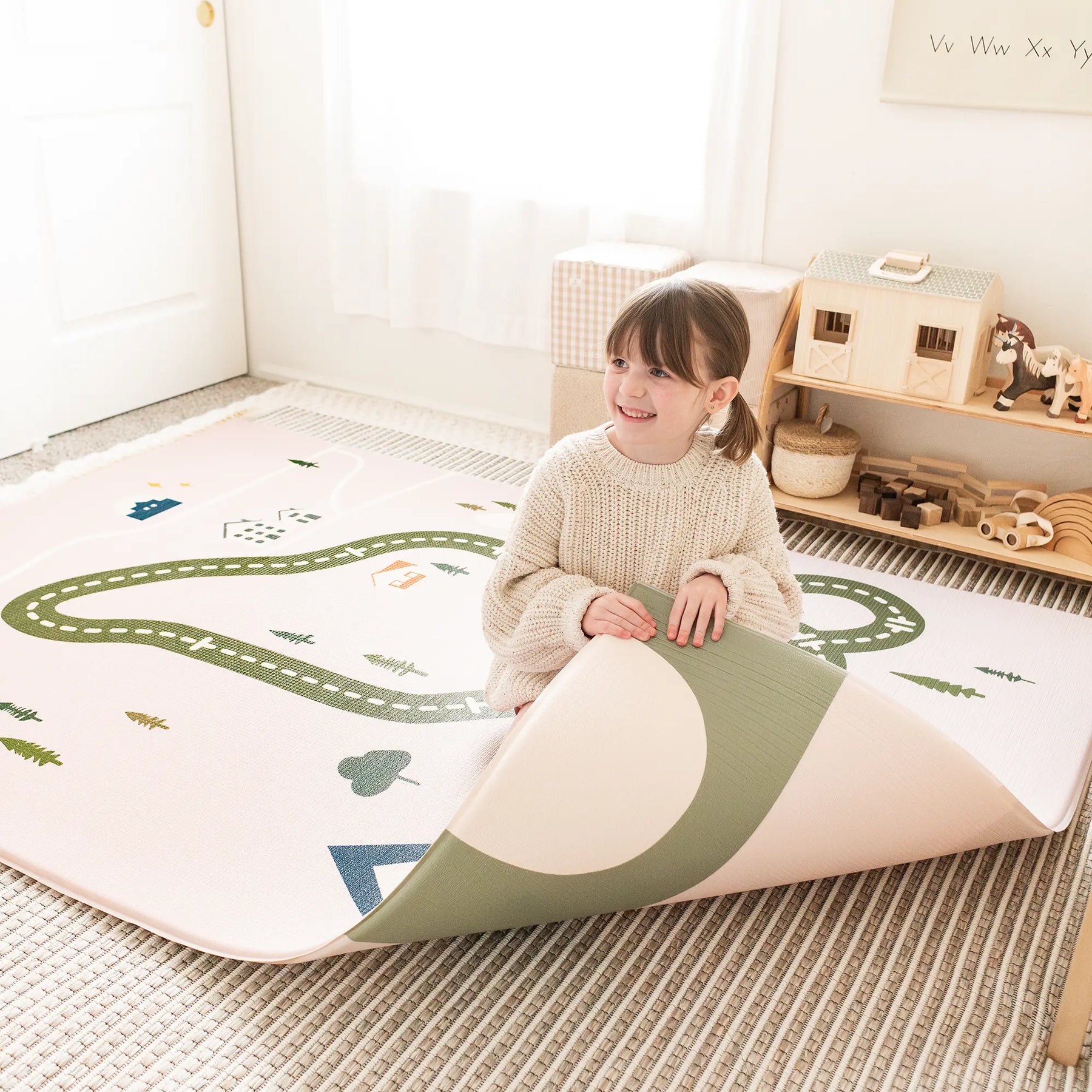 A young girl smiles as she lifts a corner of the Tiny Land® Double-Sided Baby Playmat Forest Track Wonder, made from soft GFOAM. The waterproof playmat features a road and nature design, with wooden toys and shelves in the bright room.
