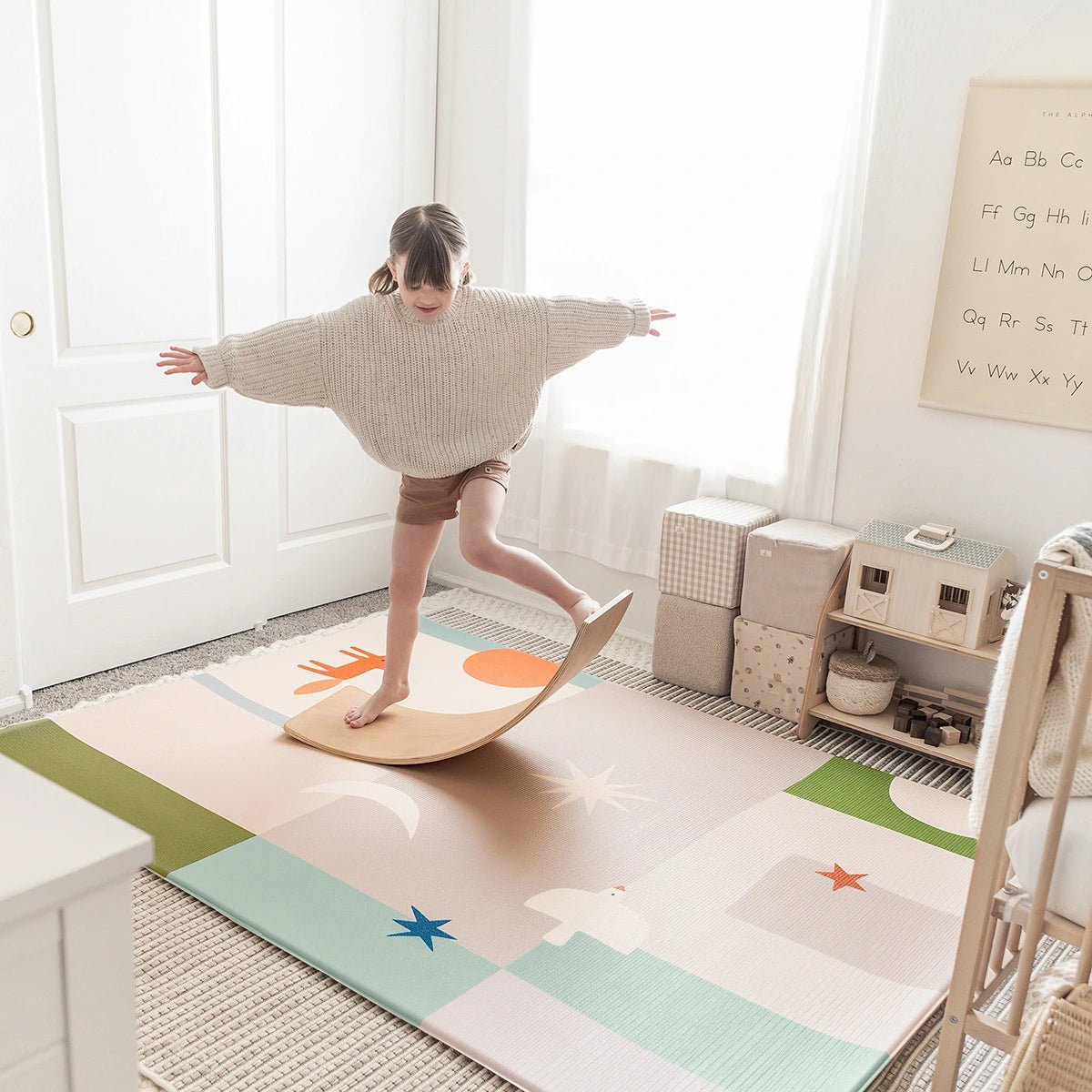 A young girl balances on a wooden board in a bright playroom featuring the Tiny Land® Double-Sided Baby Playmat Forest Track Wonder and educational wall decor.
