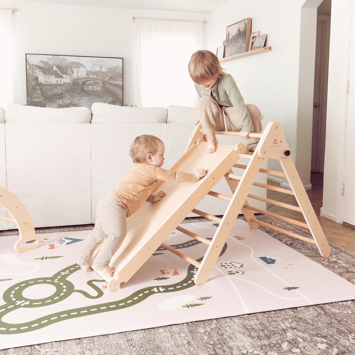 Two young children play on a wooden climbing triangle with a slide in the living room. The floor is covered by the Tiny Land® Double-Sided Baby Playmat Forest Track Wonder from Tiny Land, featuring roads and trees for imaginative fun.