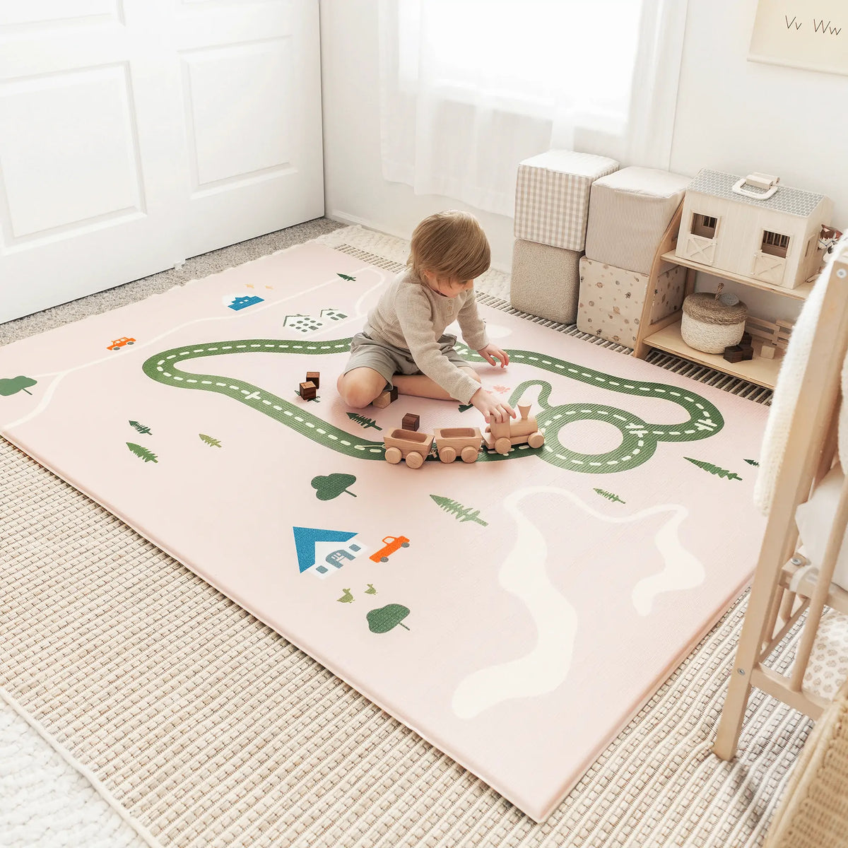 A young child sits on the Tiny Land® Double-Sided Baby Playmat Forest Track Wonder, a pink, waterproof GFOAM mat, playing with a wooden train in a bright, cozy room with pastel decor and neat storage cubes.