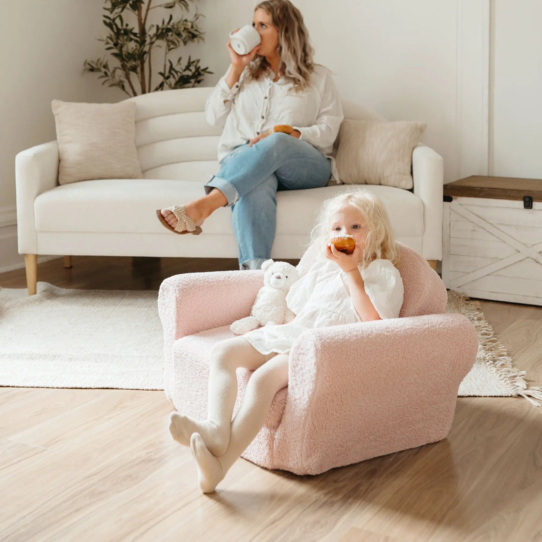A young girl enjoys a donut and holds a stuffed bear while sitting in the Tiny Land® Afternoon Couch - Pink Kids Chair by Tiny Land; behind her, a woman relaxes with a mug on a white couch in the bright, cozy living room.