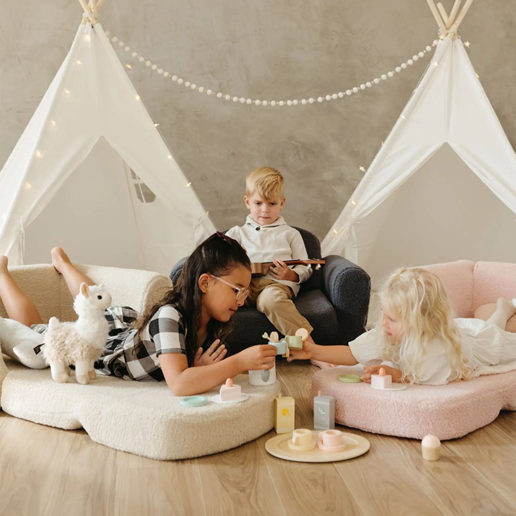 Three children play indoors near a white teepee with string lights. Two girls sit on the Tiny Land® Afternoon Couch - Pink Kids Chair by Tiny Land, surrounded by toys and a plush llama, while a boy behind them holds a book or tablet.