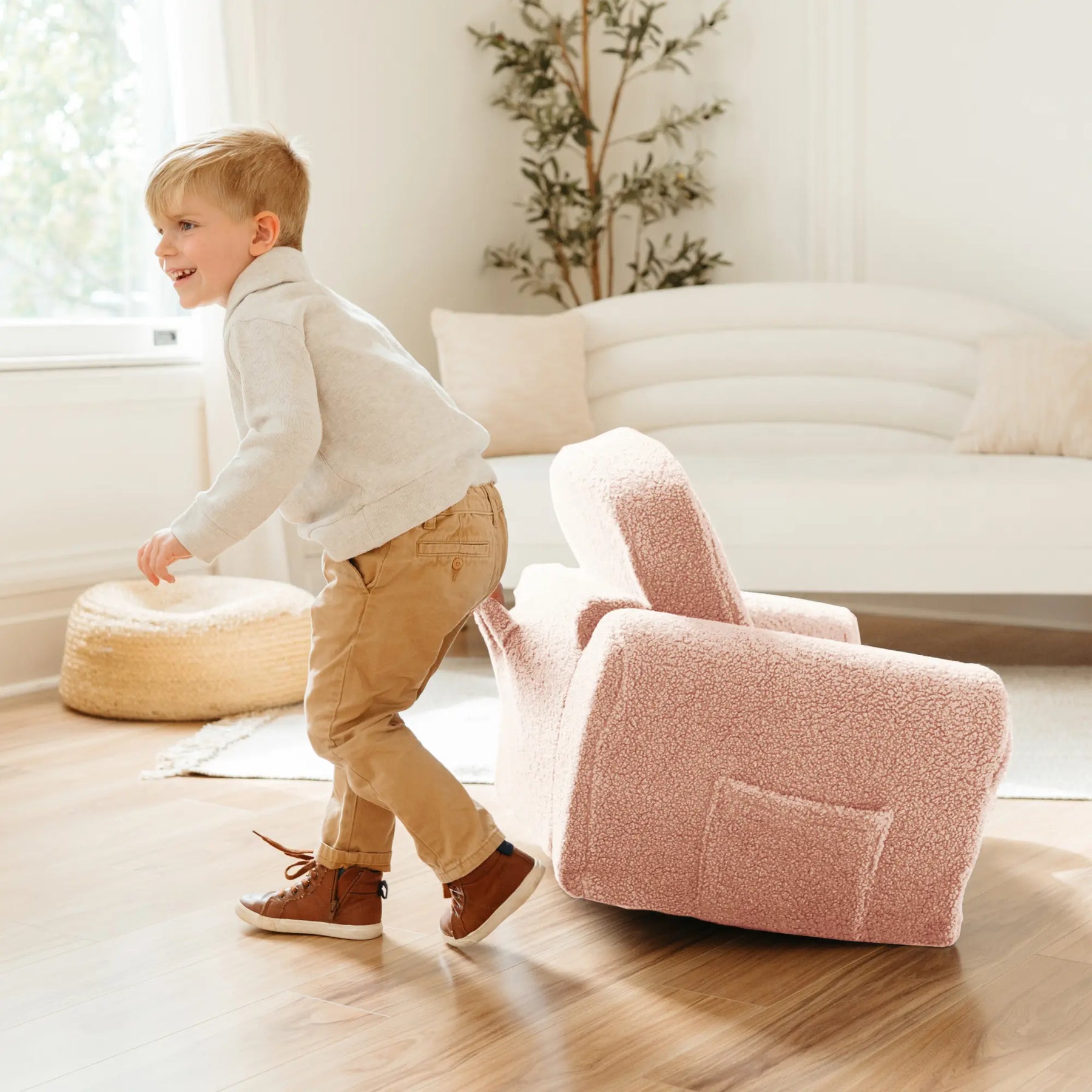 A young boy in a grey sweatshirt and tan pants smiles while playing near a tipped-over Tiny Land® Afternoon Couch - Pink Kids Chair in a bright, cozy living room.