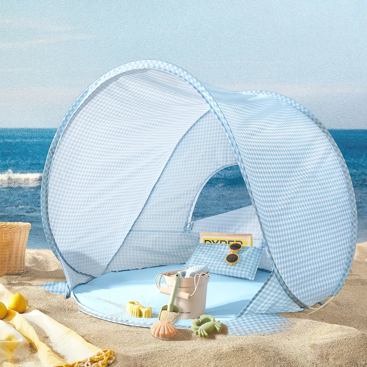 The Tiny Land® One-Stop Beach Toy & Tent Set in light blue gingham sits on a sandy beach near the ocean, with included sand play set, towel, and woven basket beside it under a clear sky.|Light Blue Gingham