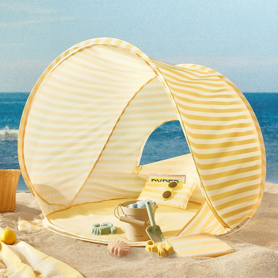 The Tiny Land® One-Stop Beach Toy & Tent Set in pastel yellow stripes sits on the sand, complete with a pillow and sand play set—bucket, shovel, and molds—by the sea under a blue sky.|Pastel Yellow Stripes