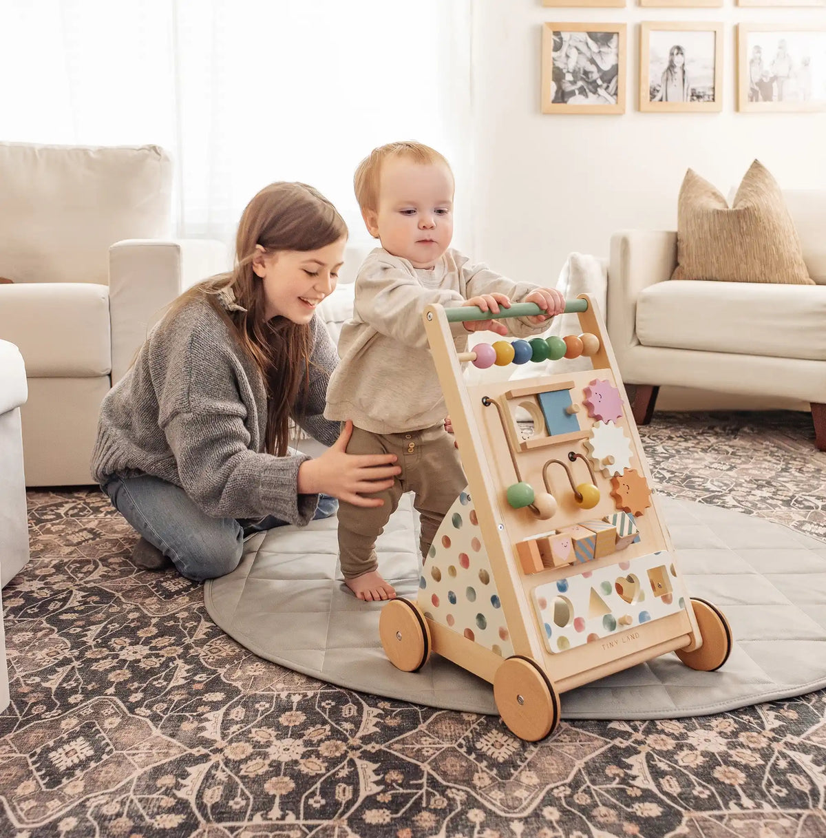 A young child stands holding the Tiny Land® Versatile Natural Wooden Baby Walker as a smiling woman kneels beside them on a patterned rug in a bright, cozy living room.