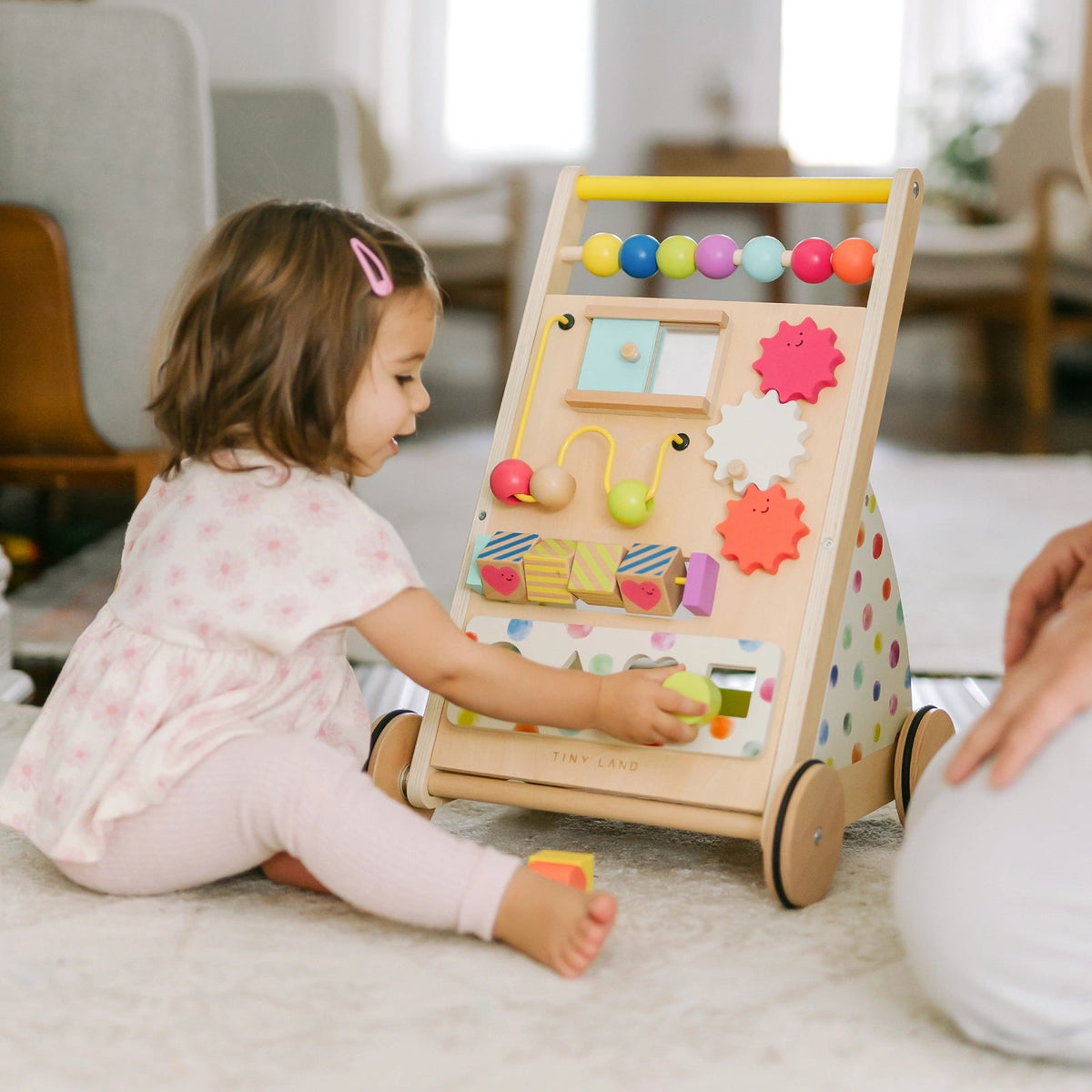 A young child in light-colored clothes plays on the floor with the Tiny Land All-Rounder Baby Playset—Baby Walker + Montessori Box (7-9 Months), filled with beads, gears, and interactive parts as an adult's hand appears nearby|Rainbow