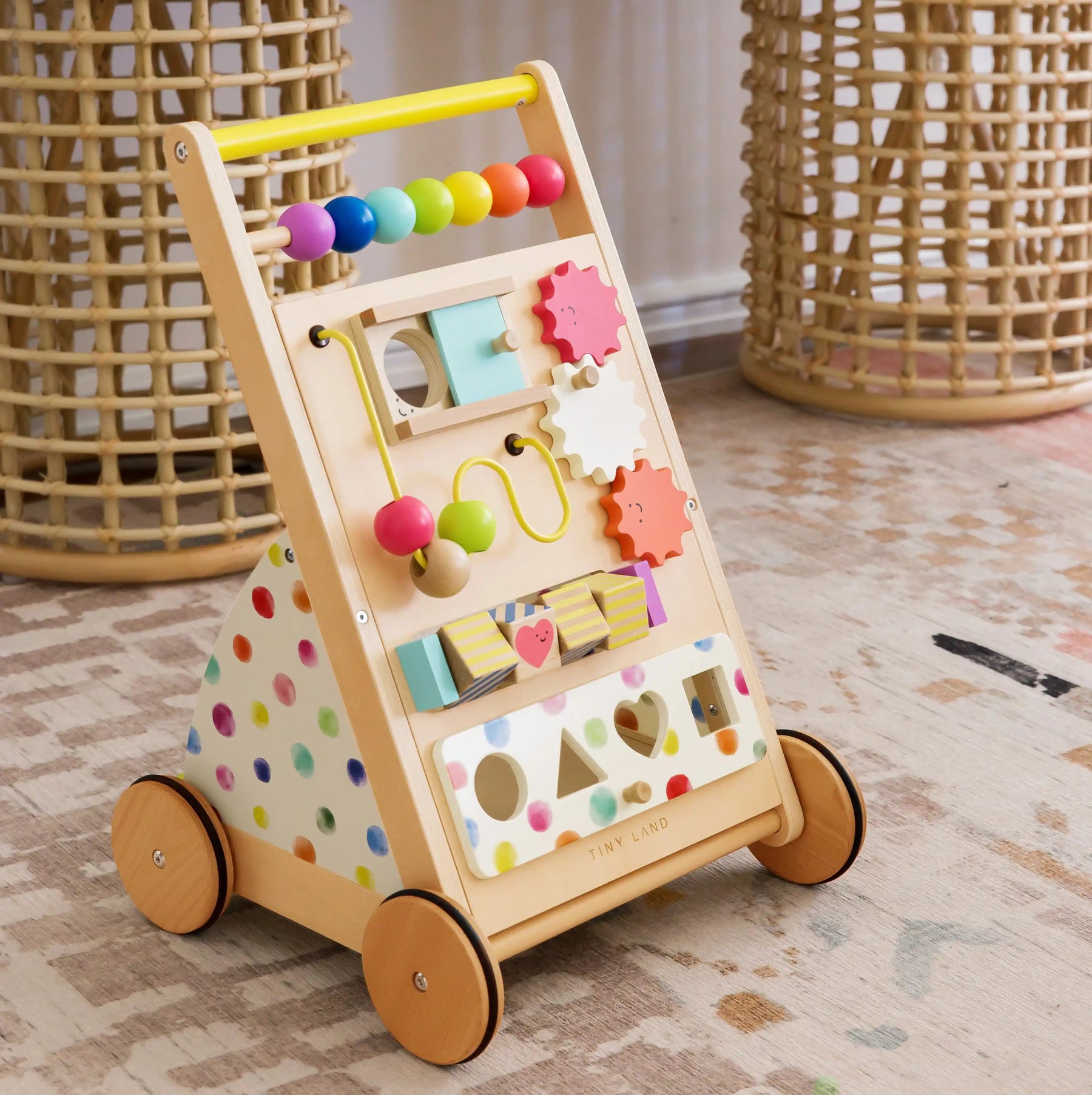 The Tiny Land® Premium Natural Wooden Activity Walker by Tiny Land, featuring colorful beads, gears, shapes, and polka dots for interactive play, sits on a patterned rug with wicker baskets in the background.