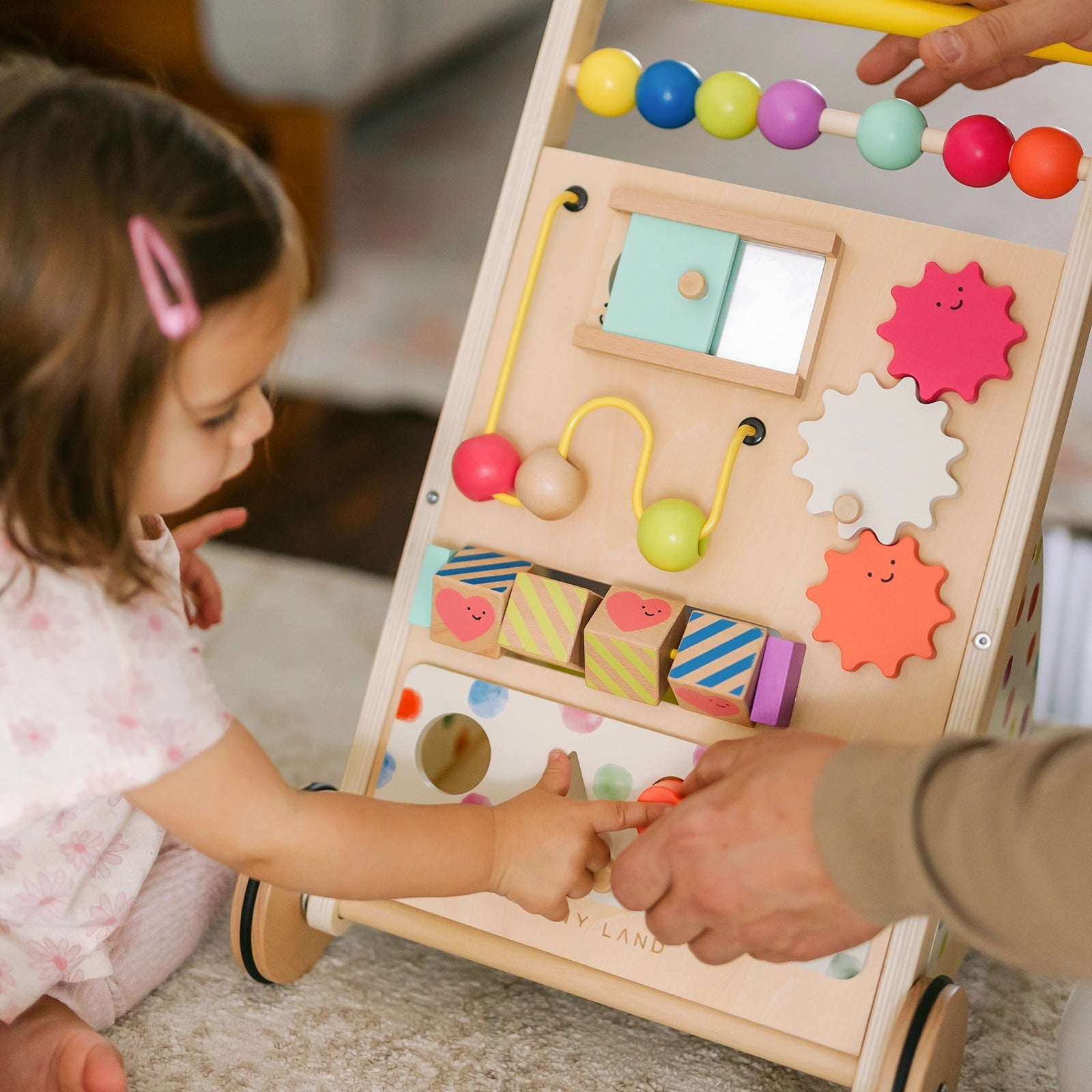 A toddler plays with the Tiny Land® Premium Natural Wooden Activity Walker, which features beads, gears, and spinning blocks, as an adult’s hand helps guide her.