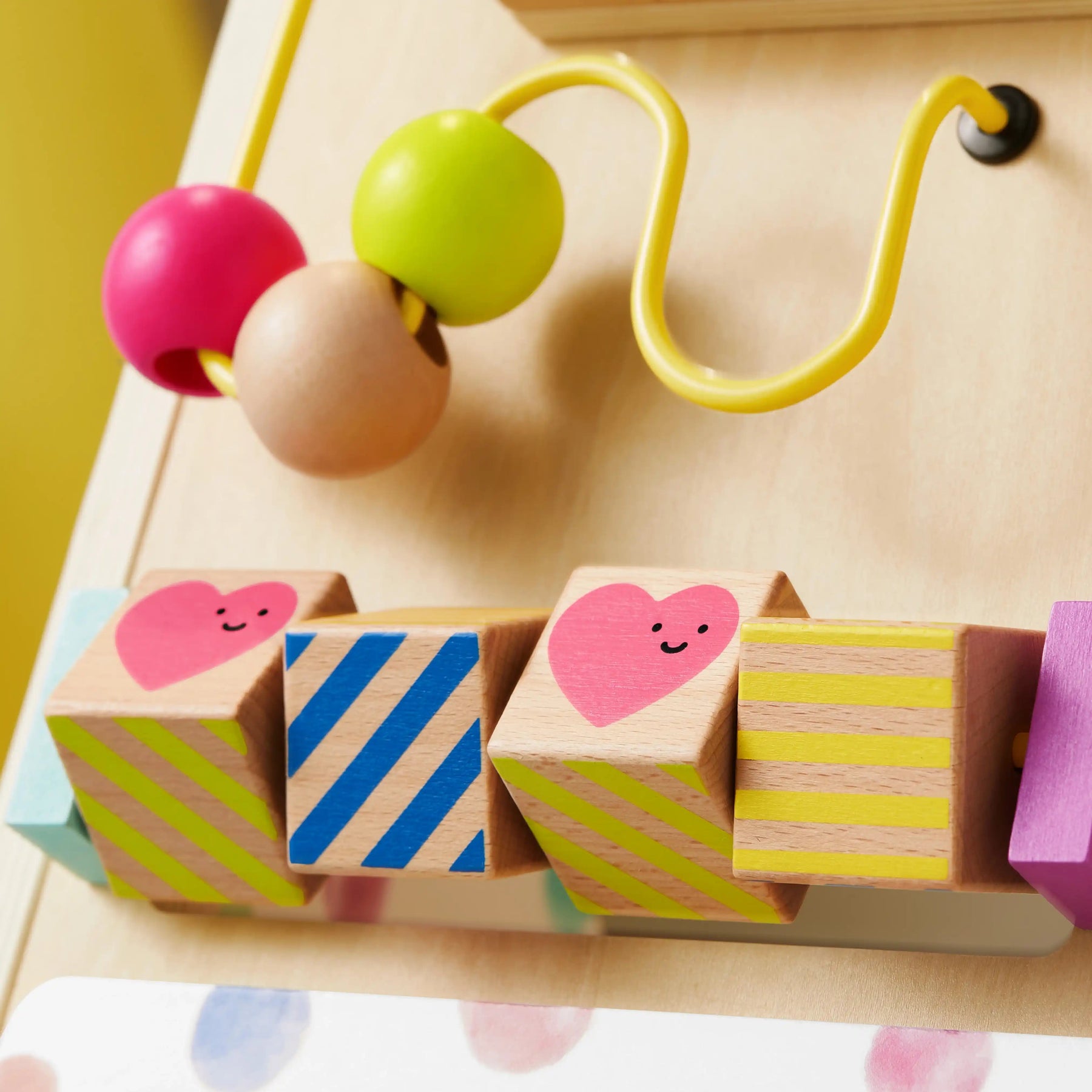 The Tiny Land® Premium Natural Wooden Activity Walker by Tiny Land features colorful beads on a yellow wire and wooden blocks with stripes and pink hearts, displayed next to a charming baby walker against a light background.