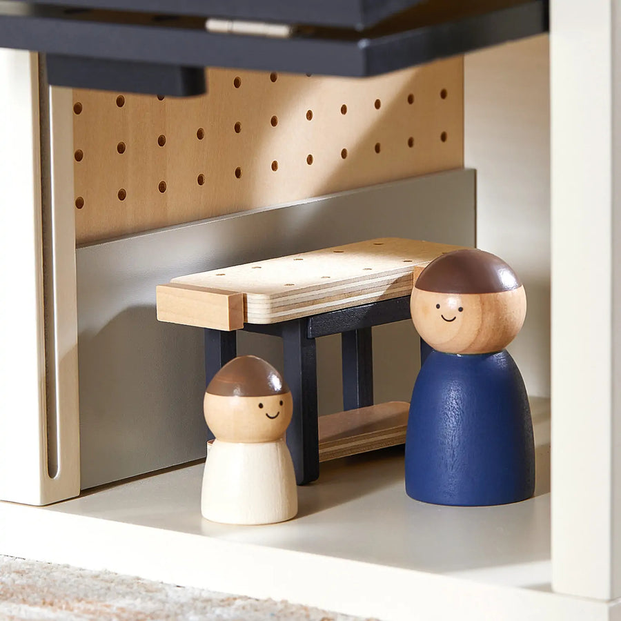 Two wooden peg dolls with brown hair stand by a workbench inside the light-colored Tiny Land® Wooden Dollhouse - Garage, which features a pegboard wall.