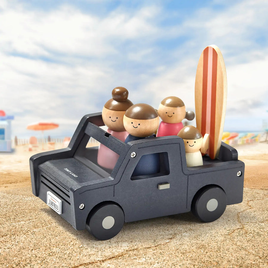 A Tiny Land® Wooden Dollhouse - Garage toy truck carries peg dolls and a striped surfboard on sand, with a blurred beach scene in the background.