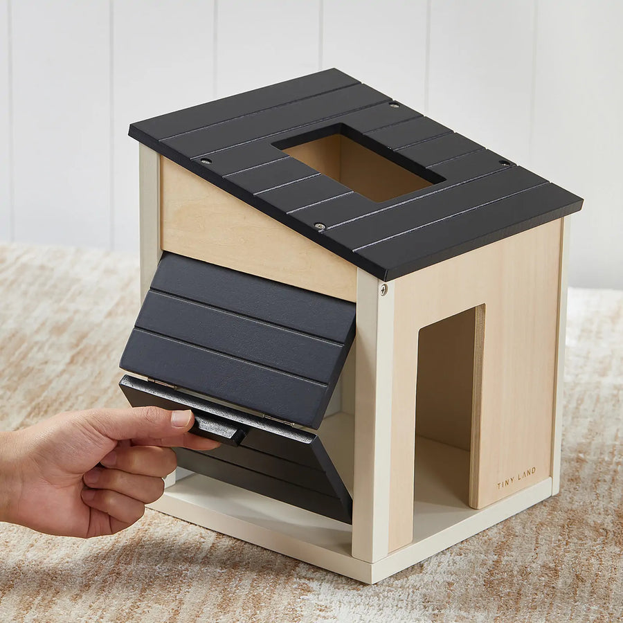 A hand lifts the black, slanted roof of the Tiny Land® Wooden Dollhouse - Garage, which features a square hole on top and an open doorway, shown on a light wood surface.