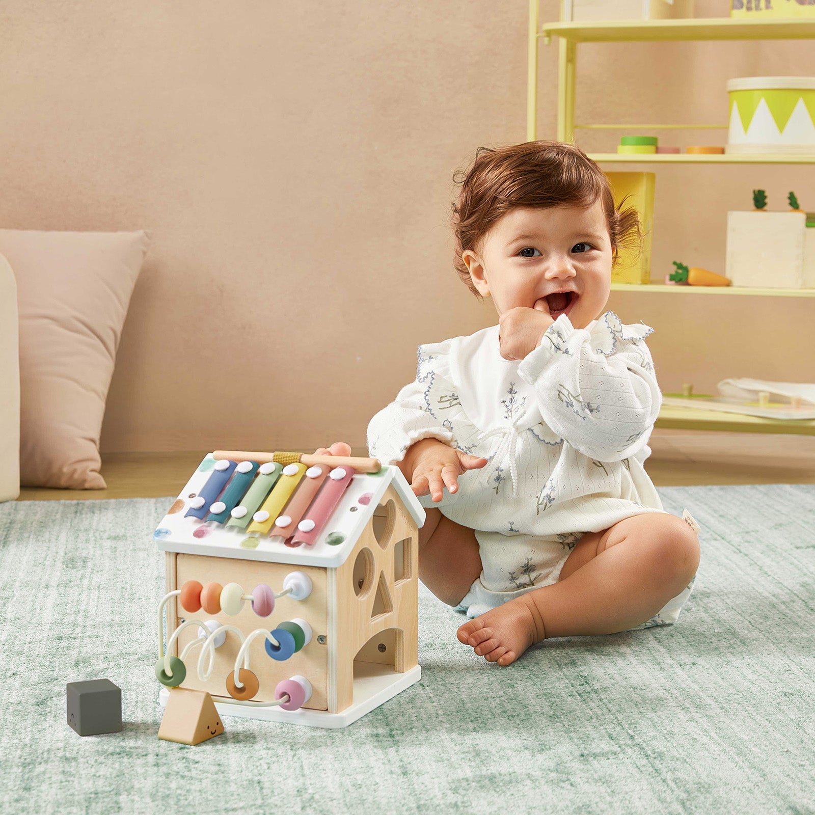 A smiling baby in a white outfit plays on the carpet with the Tiny Land® Cottage Activity Cube - Morandi by Tiny Land, shaped like a house, while shelves and toys are visible in the background.