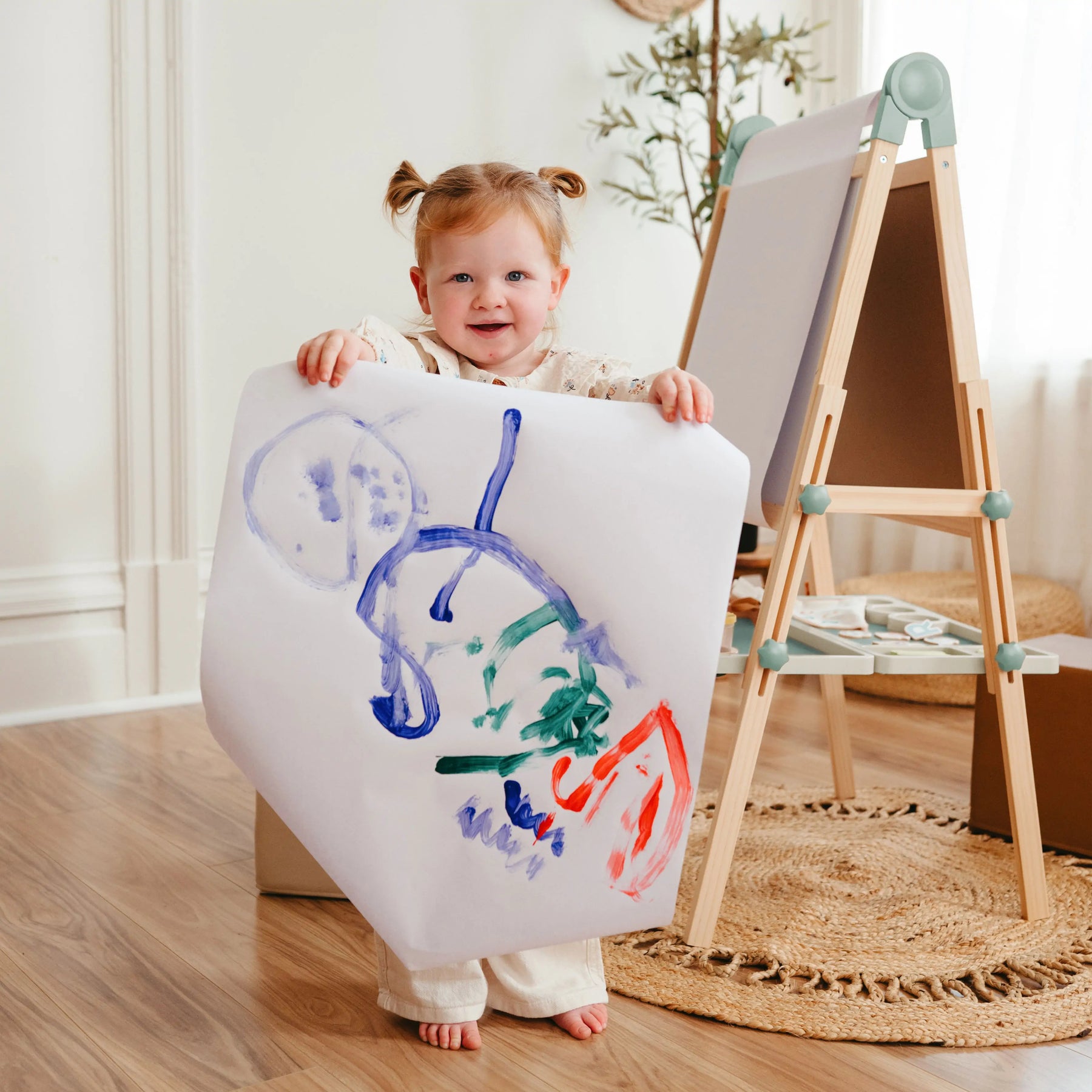A young child with pigtails proudly displays a colorful abstract painting beside her Tiny Land® Magnitales Easel for Kids - Green by Tiny Land, standing in a cozy, sunlit room with wooden floors.