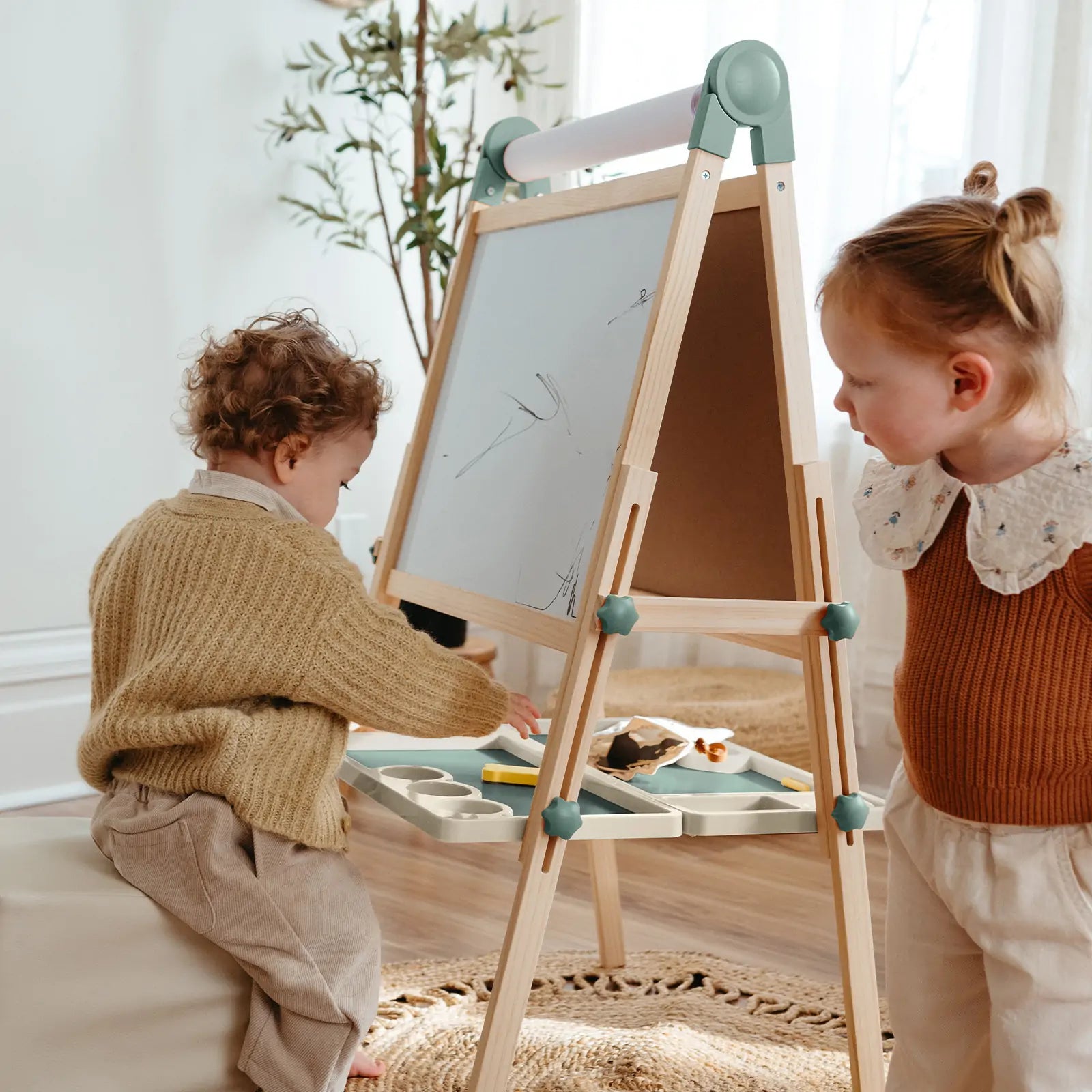 Two young children create art on the Tiny Land® Magnitales Easel for Kids - Green. One stands, one sits, both focused on their drawings. Art supplies are neatly organized in trays attached to the Tiny Land easel for easy access.
