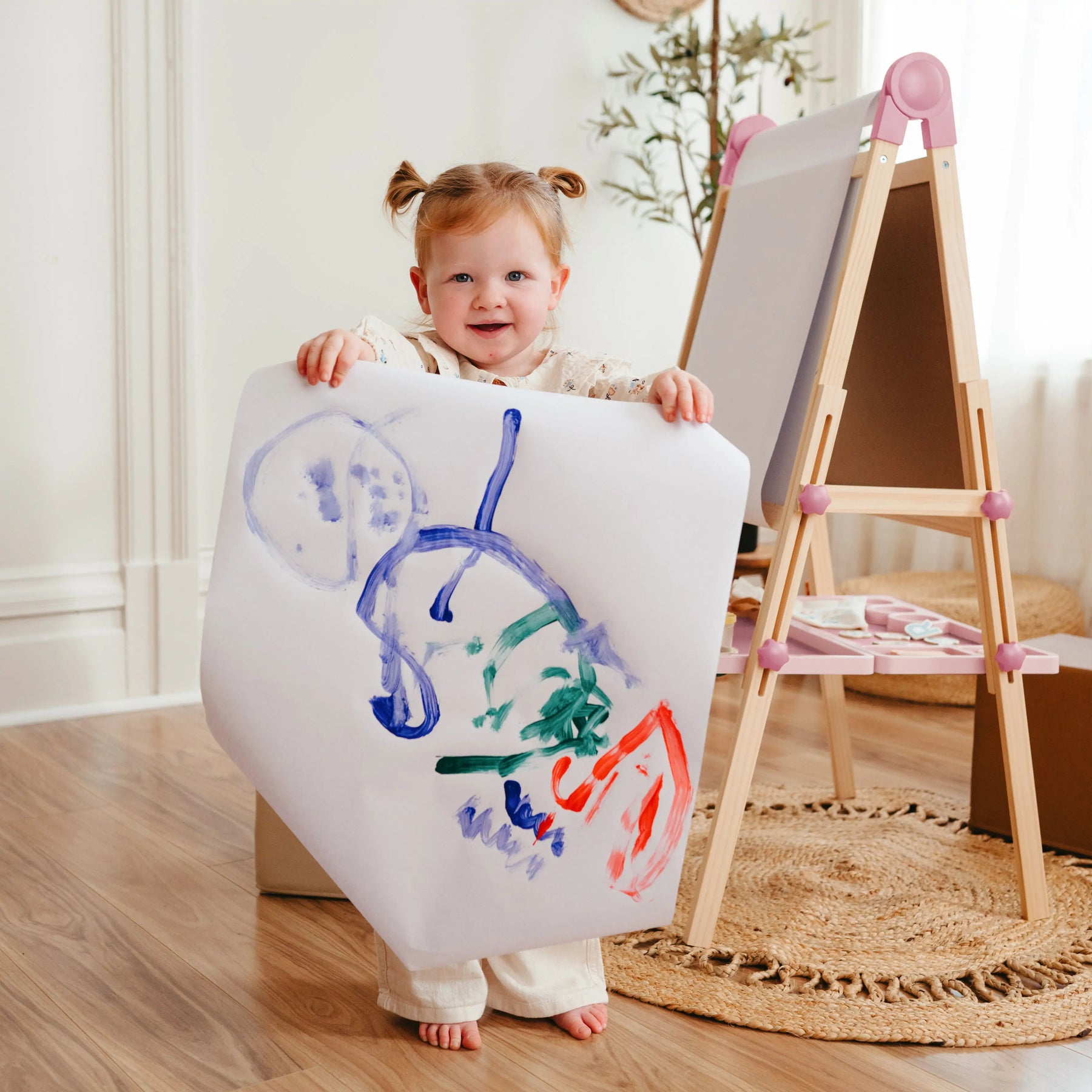A young child smiles and displays colorful abstract art while standing next to the Tiny Land® Magnitales Easel for Kids - Pink by Tiny Land in a cozy, well-lit room.