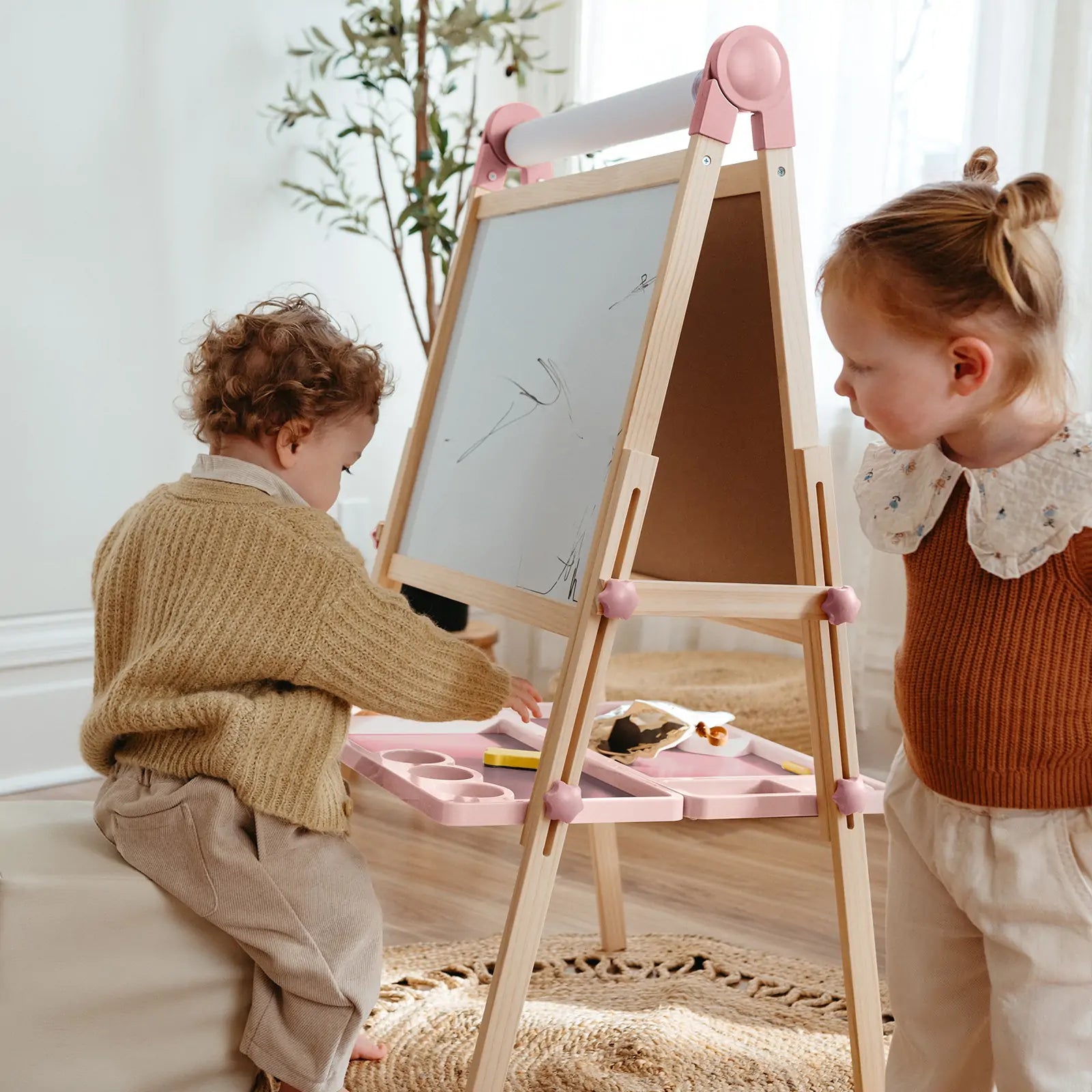 Two children draw and use art supplies together on the Tiny Land® Magnitales Easel for Kids - Pink, a height-adjustable whiteboard easel with tray, in a cozy room with sunlight, a rug, and a plant.