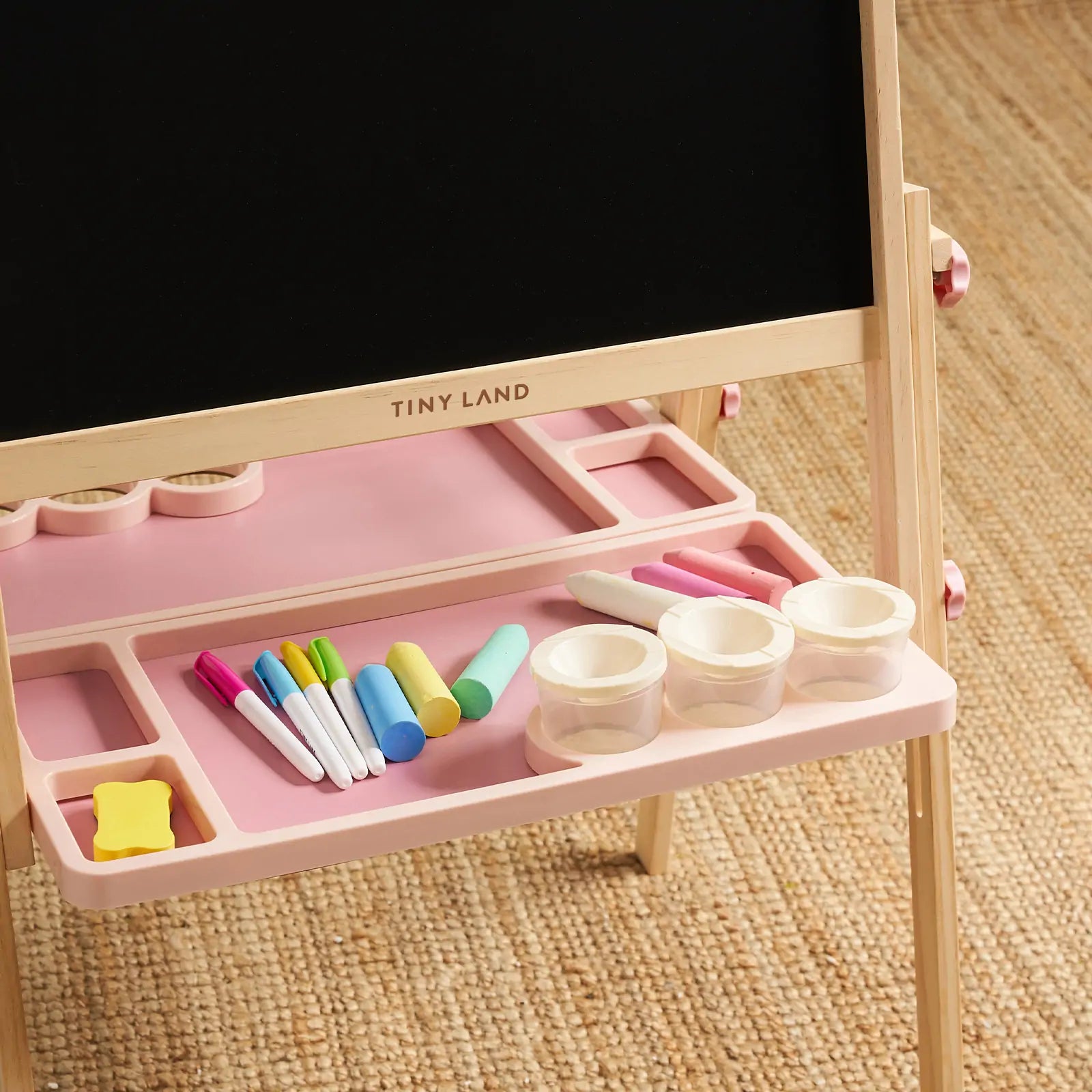 The Tiny Land® Magnitales Easel for Kids - Pink features a height adjustable blackboard, wooden legs, and tray to hold markers, chalk, a yellow eraser, and three clear cups. "Tiny Land" is printed on the wood. The easel sits on a woven carpet.