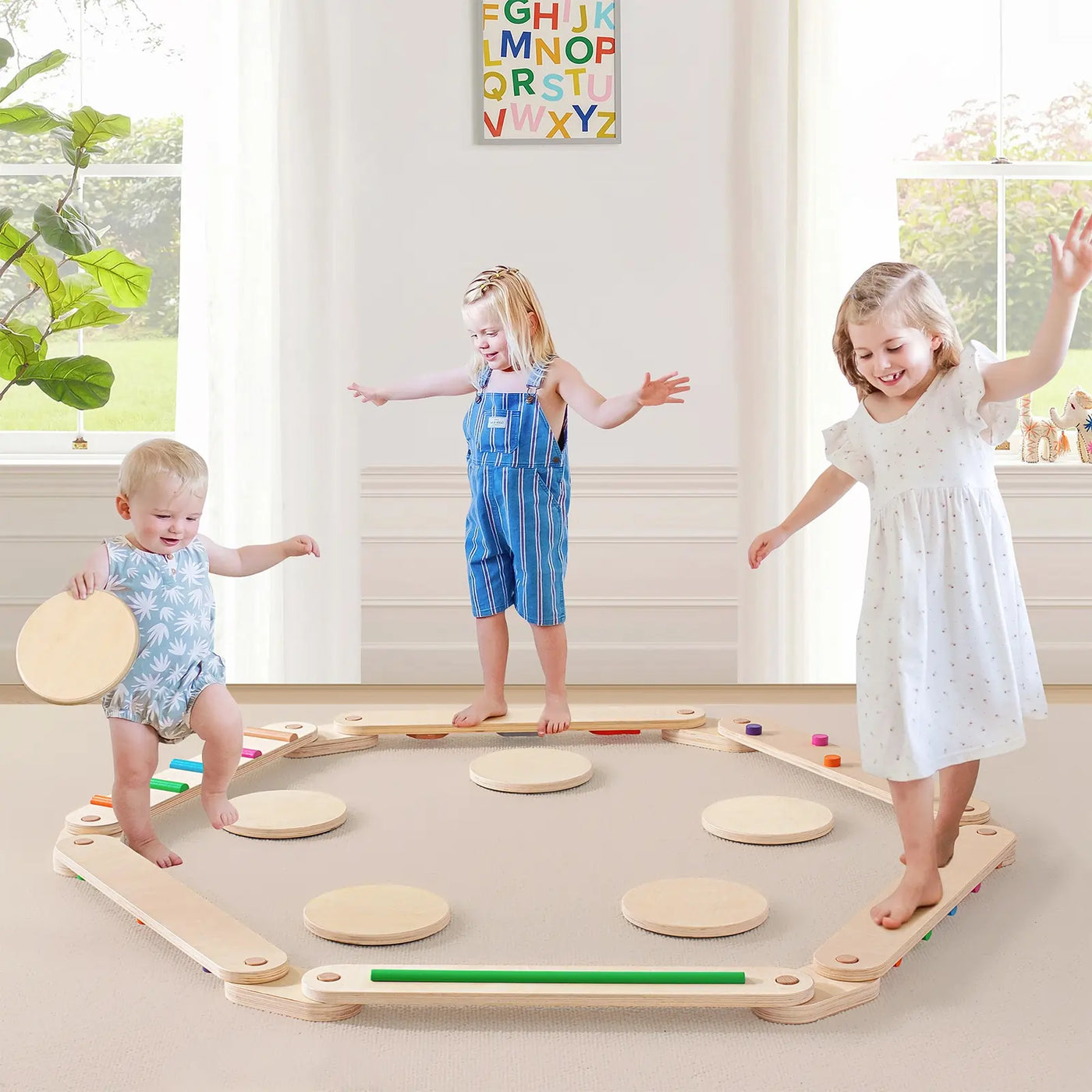 Three young children play barefoot on a wooden Tiny Land® Balance Beam obstacle course. Two girls and a toddler boy step across circular platforms, with a colorful alphabet poster brightening the sunlit room behind them.