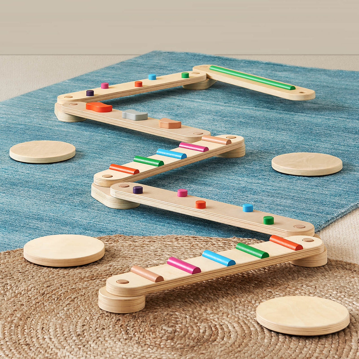 The Tiny Land® Balance Beam by Tiny Land features colorful cylindrical pegs and is arranged in a zigzag on blue and brown woven rugs, with round wooden stepping discs placed around it.