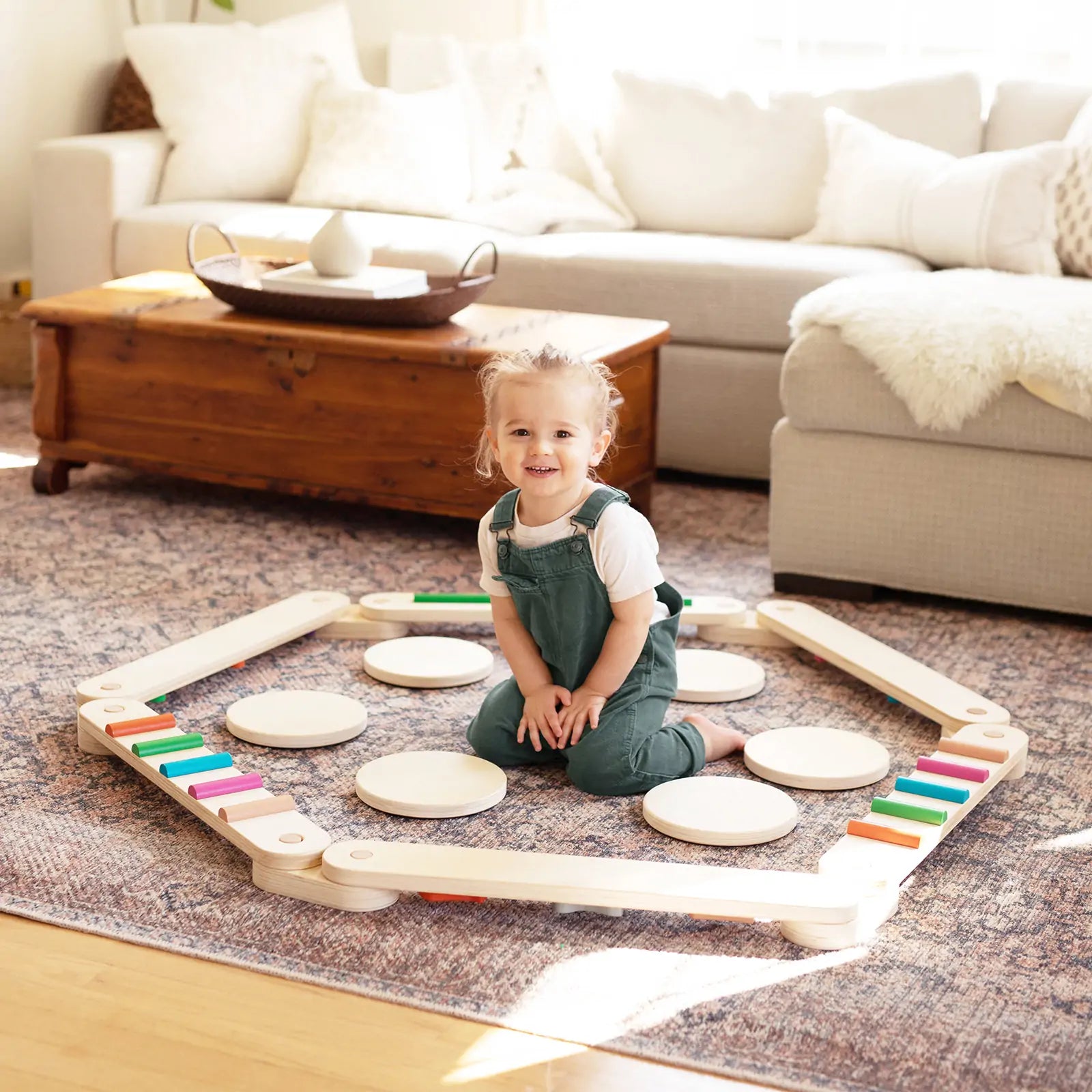 A young child in green overalls smiles on a patterned rug, surrounded by the colorful Tiny Land® Balance Beam from Tiny Land, in a cozy living room with a sofa and wooden coffee table.
