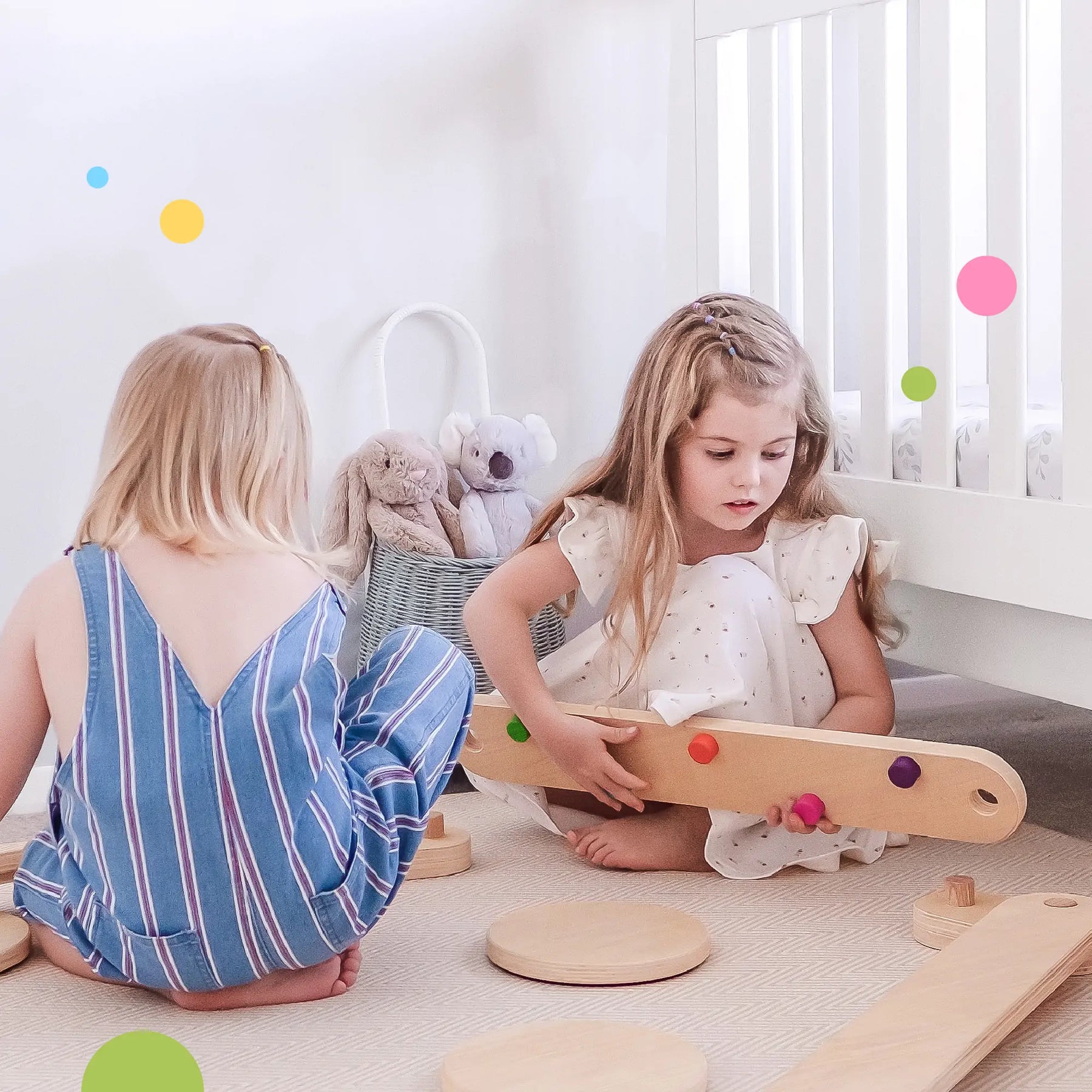 Two young girls play in a bright room with wooden toys, including the Tiny Land® Balance Beam by Tiny Land. One wears a blue striped dress and the other a white dress. A crib, stuffed animals, and pastel dots decorate the background.