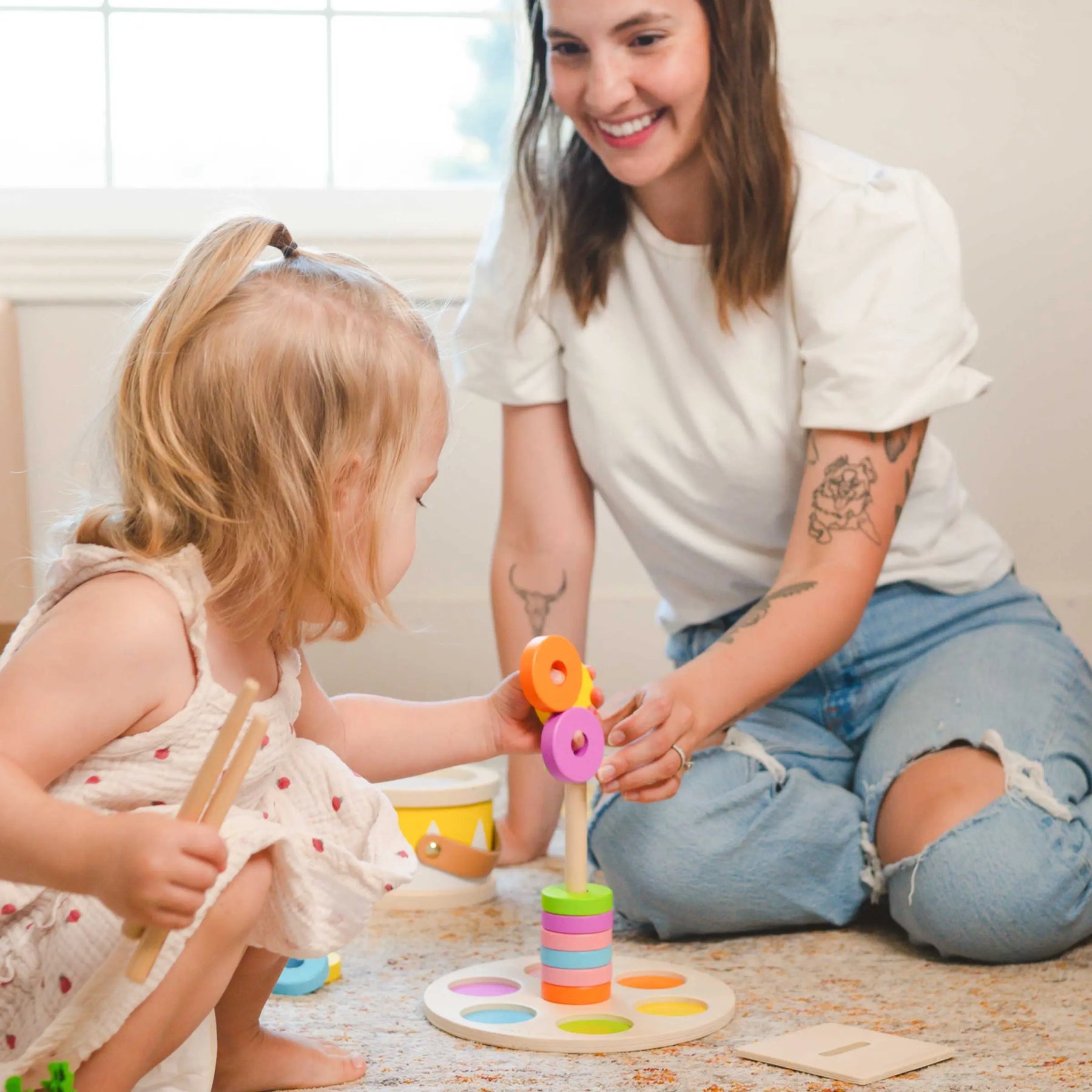 A woman in a white shirt and ripped jeans smiles as she plays with a 13–15 month toddler, who explores the Tiny Land® Montessori Toys for Toddlers and practices stacking rings from the Tiny Land learning box.