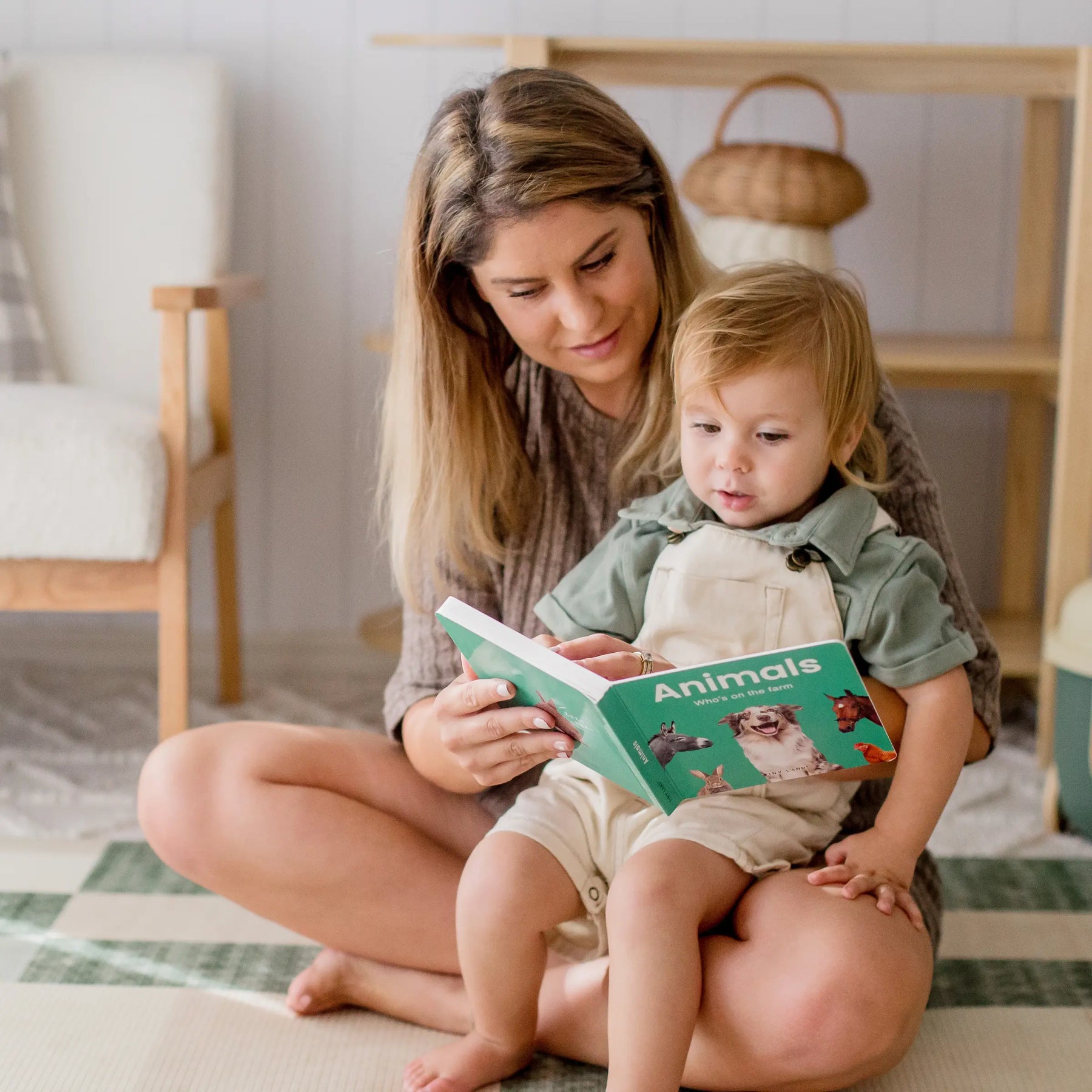 A woman reads a colorful book with a young child in her lap beside the Tiny Land® Montessori Toys for Toddlers (16–18 Months)—educational toys by Tiny Land, perfect for sparking curiosity in a cozy, softly lit room.