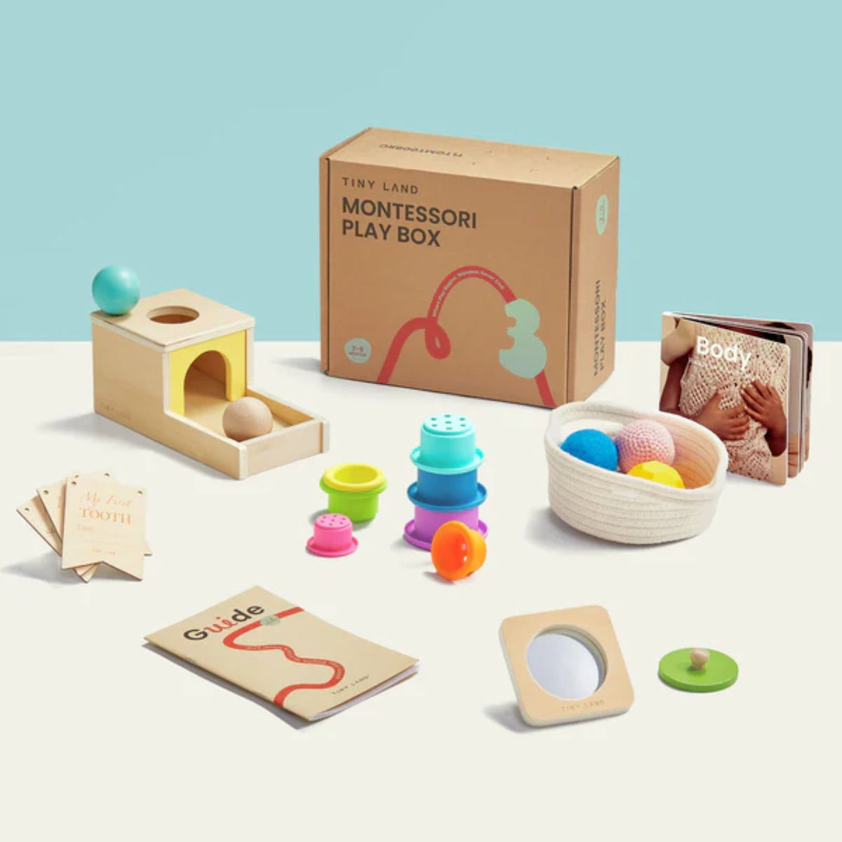 The Tiny Land® Montessori Complete Baby Set (0-24 months) by Tiny Land features wooden toys, colorful stacking cups, sensory balls in a basket, the book "Body," cards, and a guide—all displayed on a light blue and beige background.