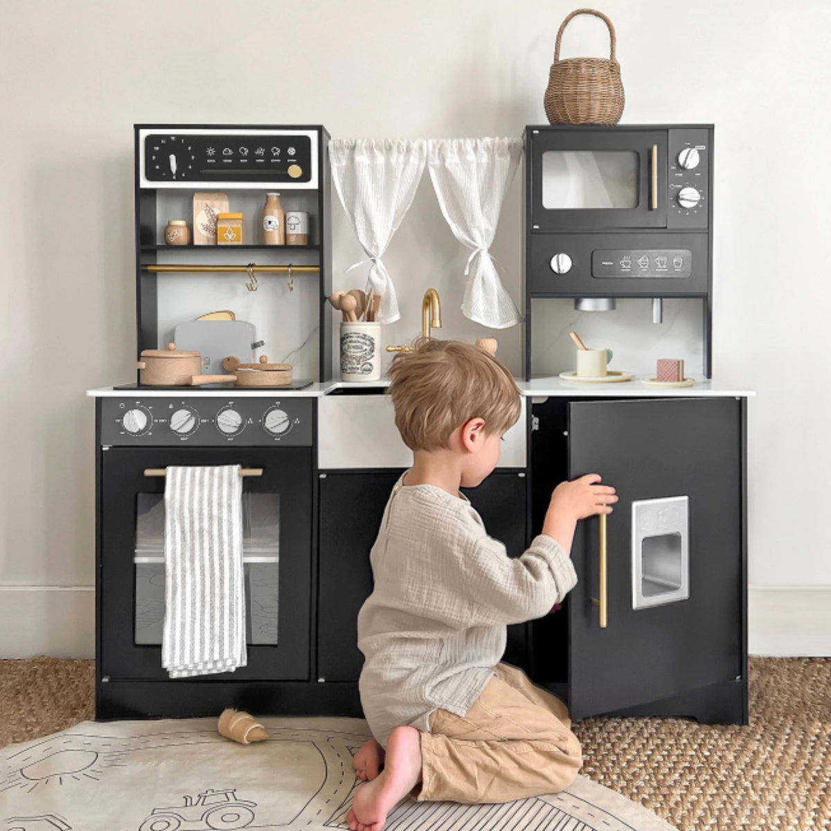 A young child plays on the floor with the Tiny Land® Iconic Wooden Play Kitchen – Black, featuring an oven, stove, sink, microwave, utensils, pots, fabric curtains, and is opening a cabinet door.