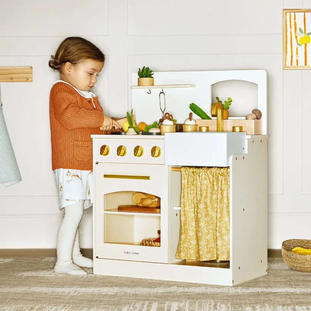 A young child stands beside the Tiny Land® Chef’s Corner Playset – Cream Playhouse + Cream Kitchen Set, pretending to cook with toy vegetables and accessories. The set features gold-colored knobs and a yellow patterned curtain in a softly lit room.