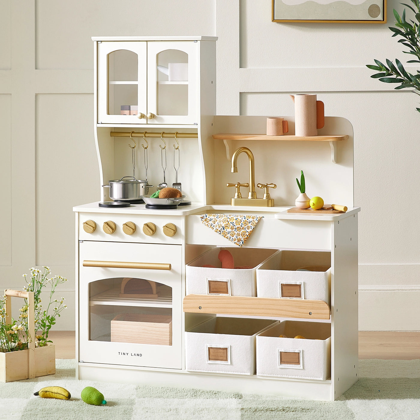 The Tiny Land® Wooden Play Kitchen with Storage by Tiny Land features a stove, oven, sink with golden faucet, hanging utensils, and six white bins. Pretend cookware and toy food are arranged on the counter and floor in a bright, cozy room.