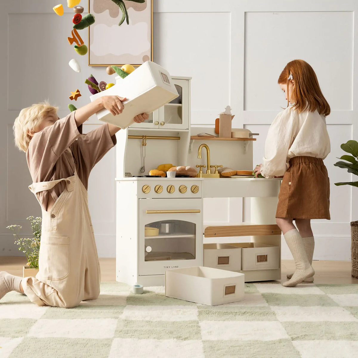 Two children play with the Tiny Land® Wooden Play Kitchen with Storage for Creative Toddlers by Tiny Land; one kneels, tossing toy vegetables from a box, while the other watches in a bright room with a soft green and white checkered rug.
