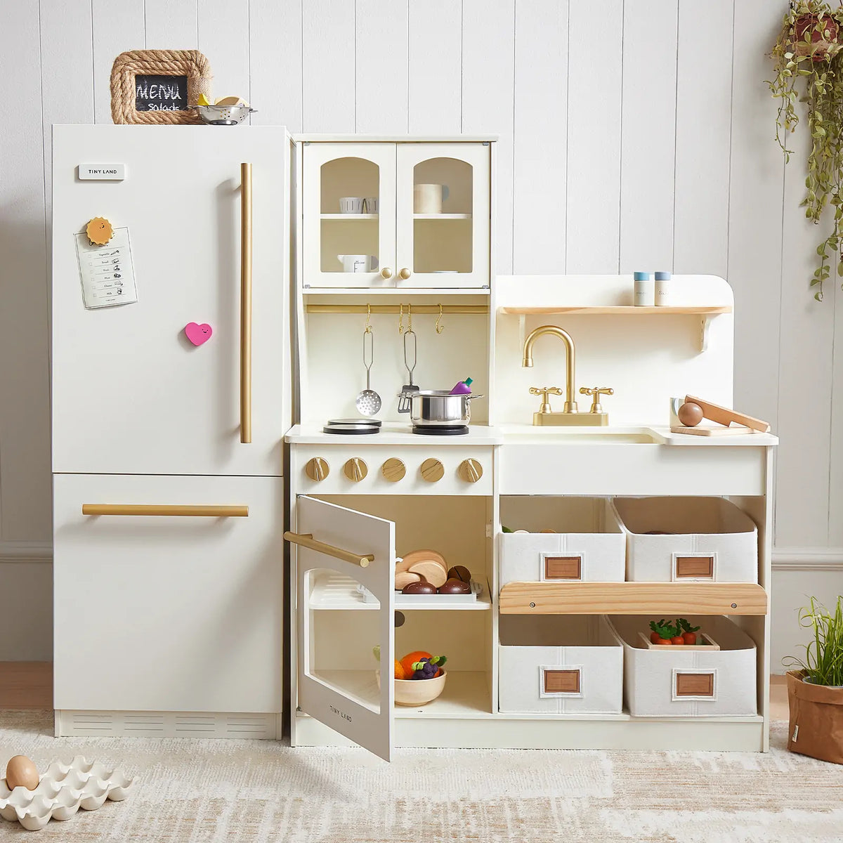 The Tiny Land Food-Filled Kitchen Gift Set features a storage kitchen, fridge, and organic food collection in white and light wood tones—complete with toy stove, oven, sink, shelves, pots, bins, and toy food.