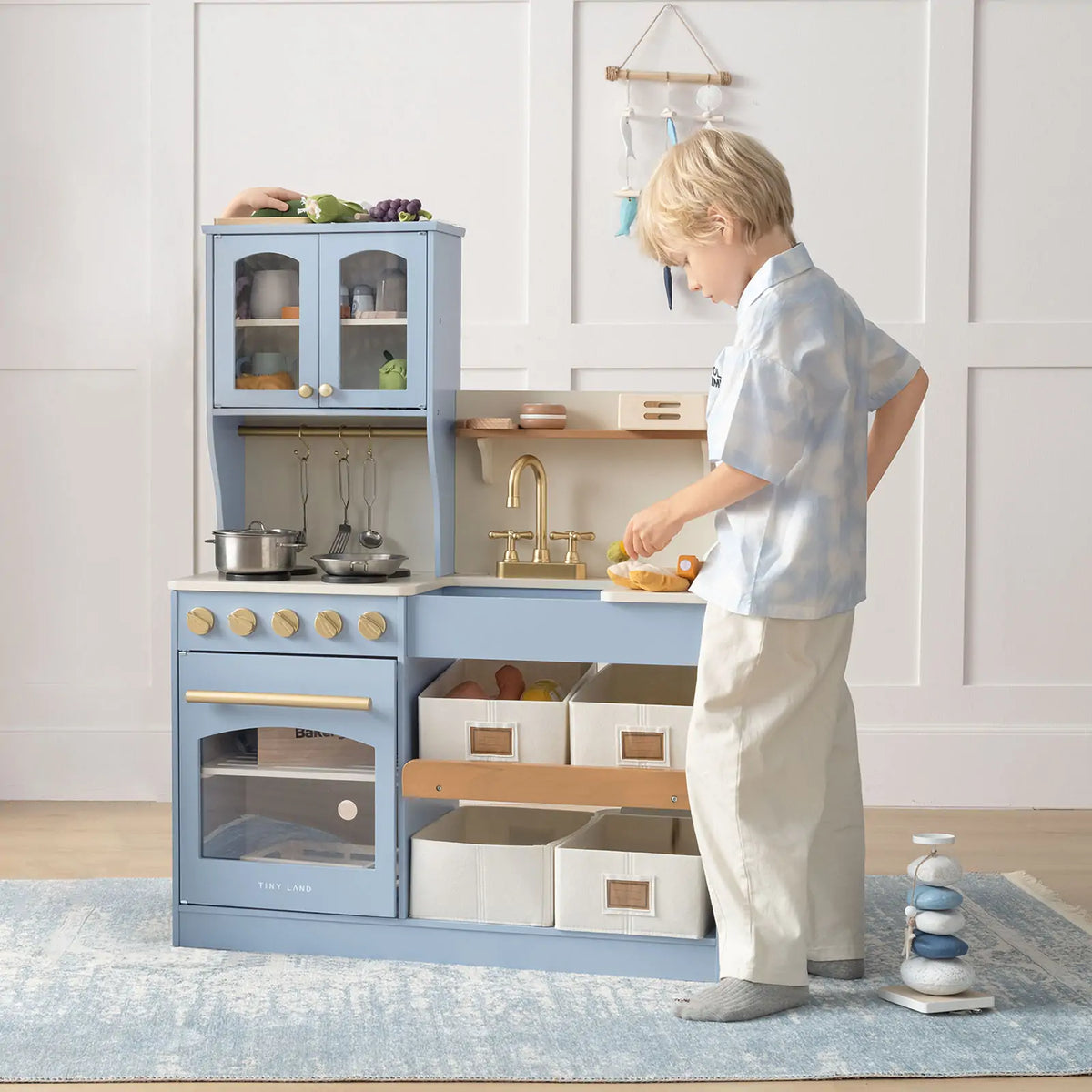 A young boy plays with the Tiny Land® Pretend Play Kitchen for Kids - Serenity Wooden Kitchen in blue, using shelves, a sink, stove, storage bins, toy cookware, and food for imaginative fun in a bright and cozy scene|blue