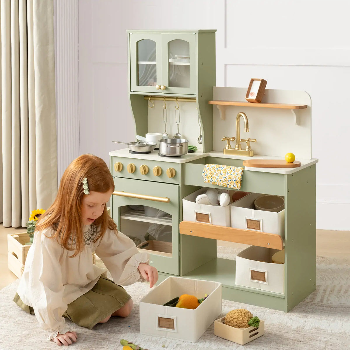 A young red-haired child plays on the floor near the Tiny Land Little Green World Gift Set: Sage Dollhouse & Sage Storage Kitchen with storage bins, toy food, and play cookware in a bright room—perfect for hours of imaginative play|green