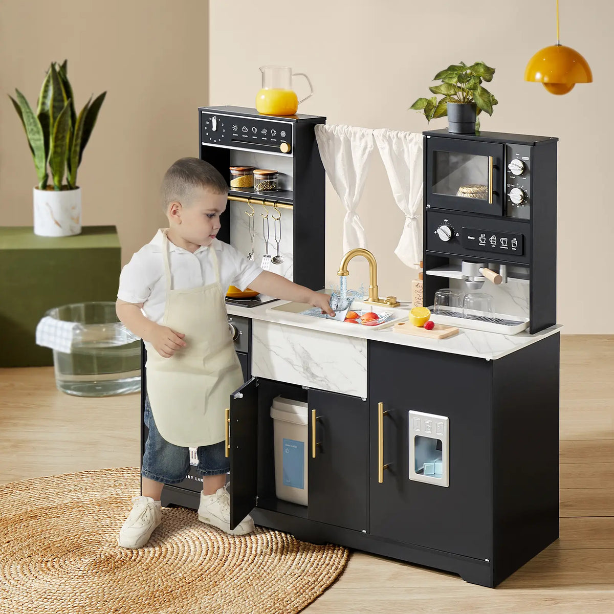A young child in an apron plays with the Tiny Land® Play Kitchen with Running Water - Iconic Aqua Kitchen in Black, featuring a Real-Flow Water System, sink, oven, and stove. A green plant and basket sit behind on a round woven rug.