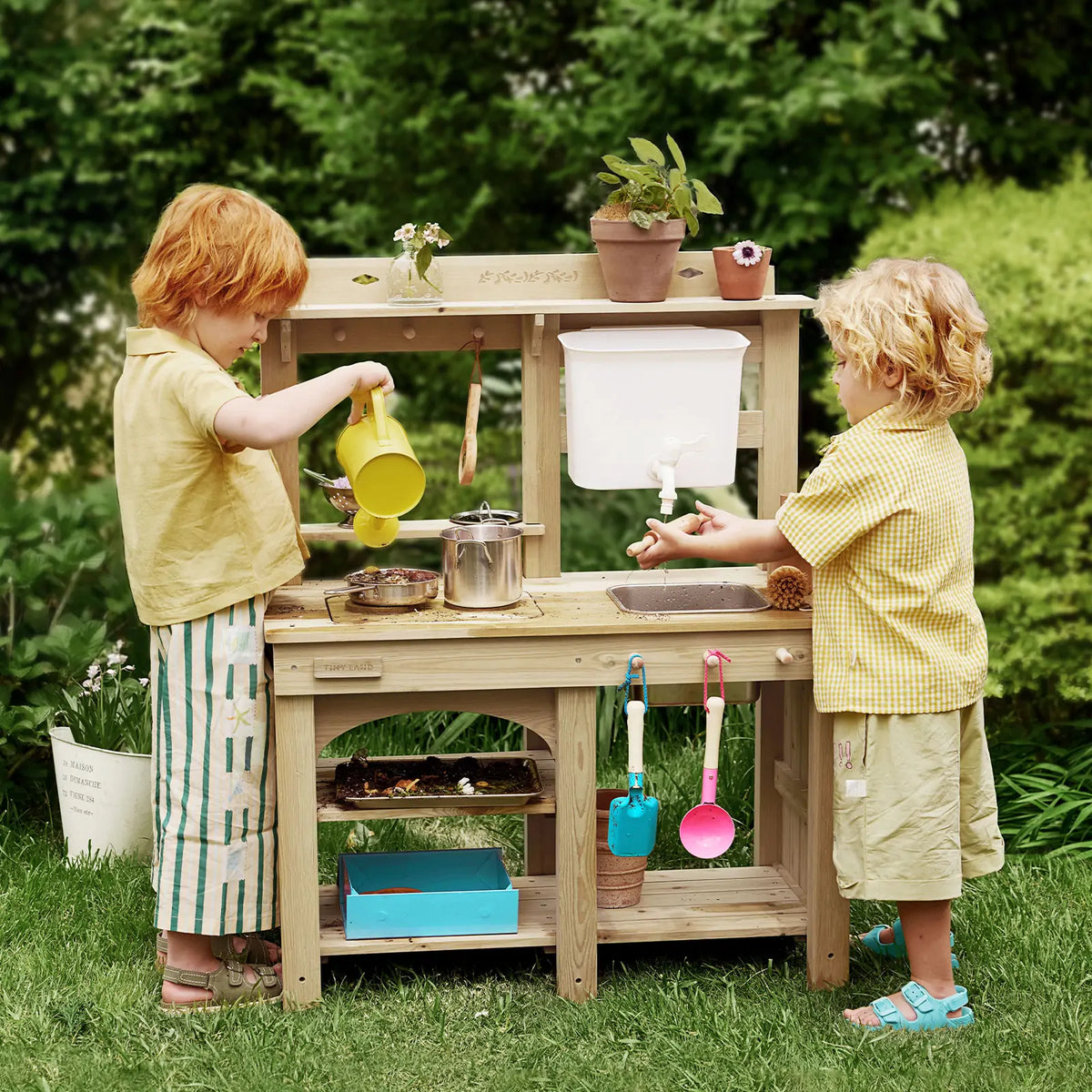 Two kids enjoy sensory play at the Tiny Land® Mud Kitchen - DuoPlay Outdoor Play Kitchen for Kids, using pots, utensils, and potted plants outdoors on green grass with trees, for endless imaginative fun by Tiny Land.
