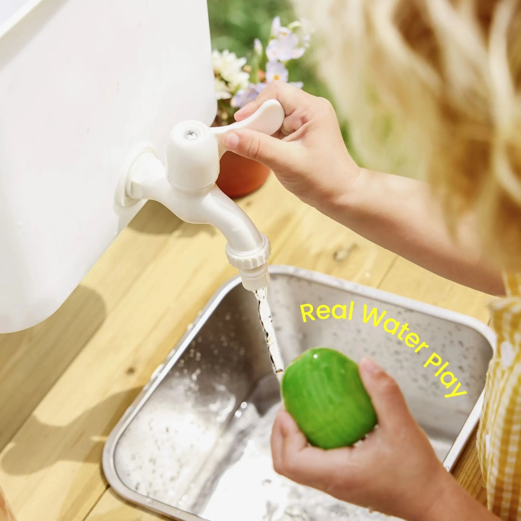 A child enjoys sensory play with the Tiny Land® Mud Kitchen - DuoPlay Outdoor Play Kitchen for Kids, holding a green sponge under running water from a white faucet over a metal sink on wood. "Real Water Play" appears in yellow beside the sink.
