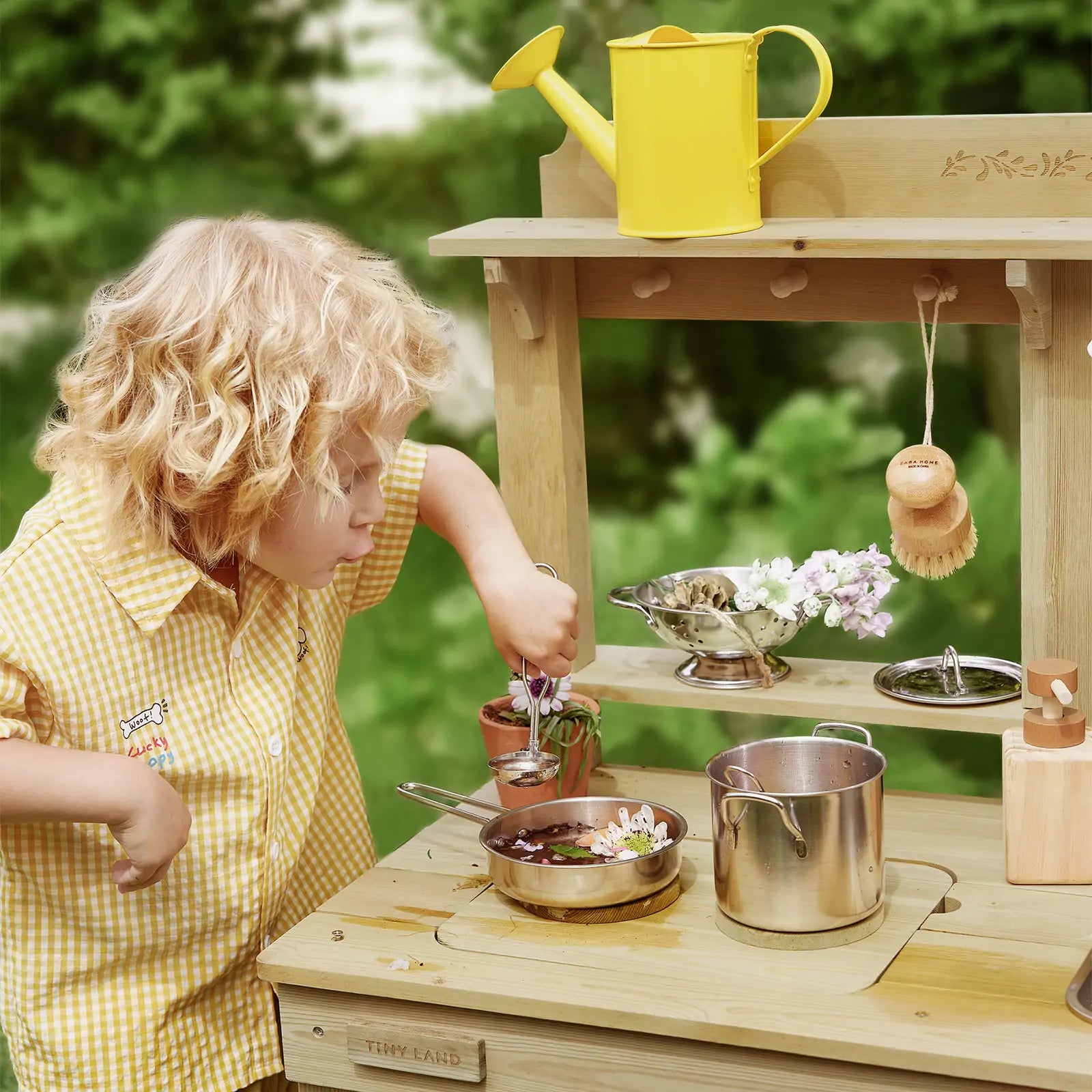 A young child in a yellow checked shirt enjoys sensory play with the Tiny Land® Mud Kitchen - DuoPlay Outdoor Play Kitchen for Kids, stirring a pot among metal cookware, flowers, and a yellow watering can. Lush greenery is blurred in the background.