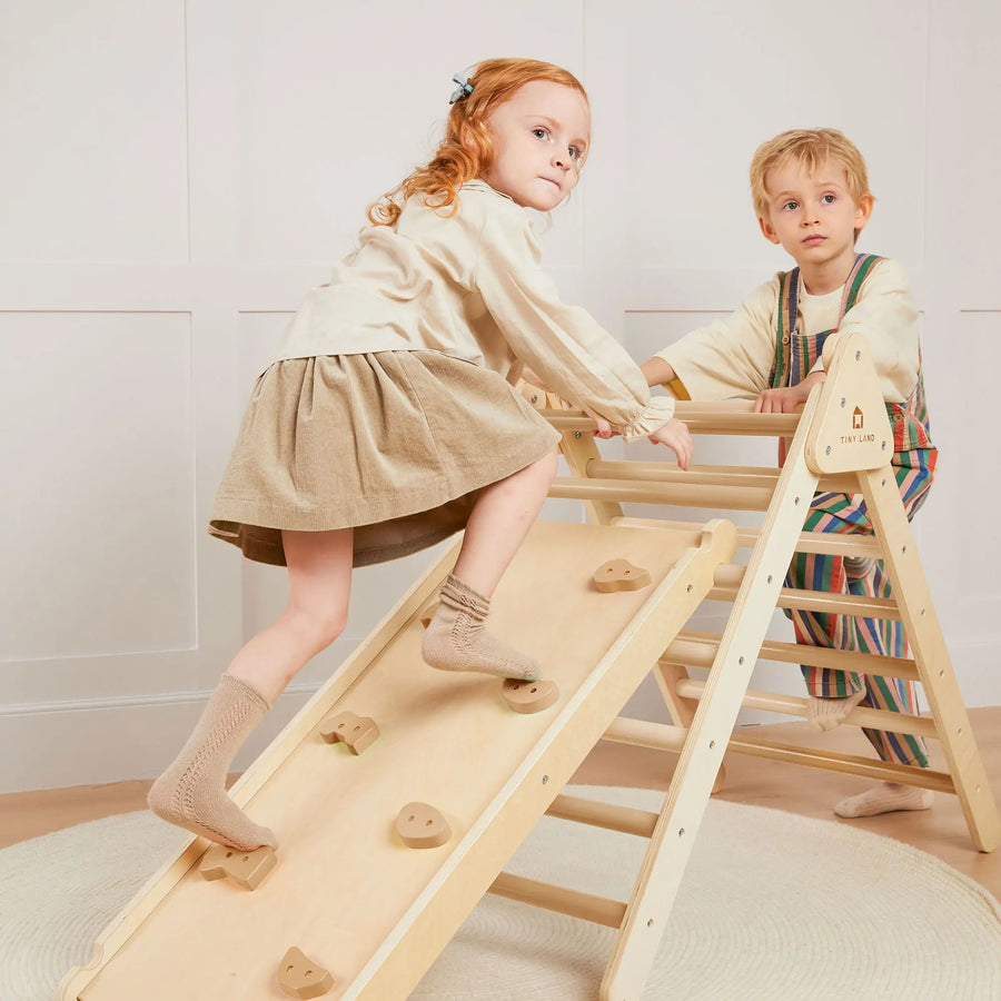 Two young children play in a bright room with the Tiny Land® Pikler Triangle 7-in-1 Montessori Climbing Set. The girl climbs the ramp while the boy stands on the other side, both engaged with the Tiny Land climbing toy.|natural wood color
