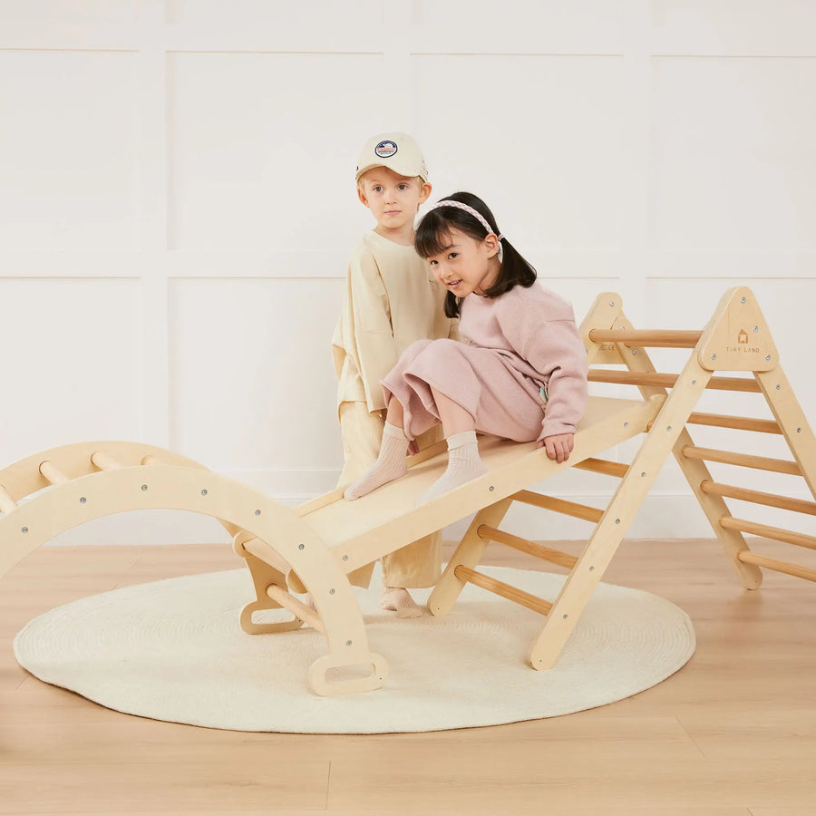 Two young children play in a bright room with the Tiny Land Safe Explorers Playset—Natural Wood Climbing Set and Urban Zoo Playmat. The girl in pink sits on the slide, while the boy in beige stands nearby on the round, cream-colored mat.|natural wood color